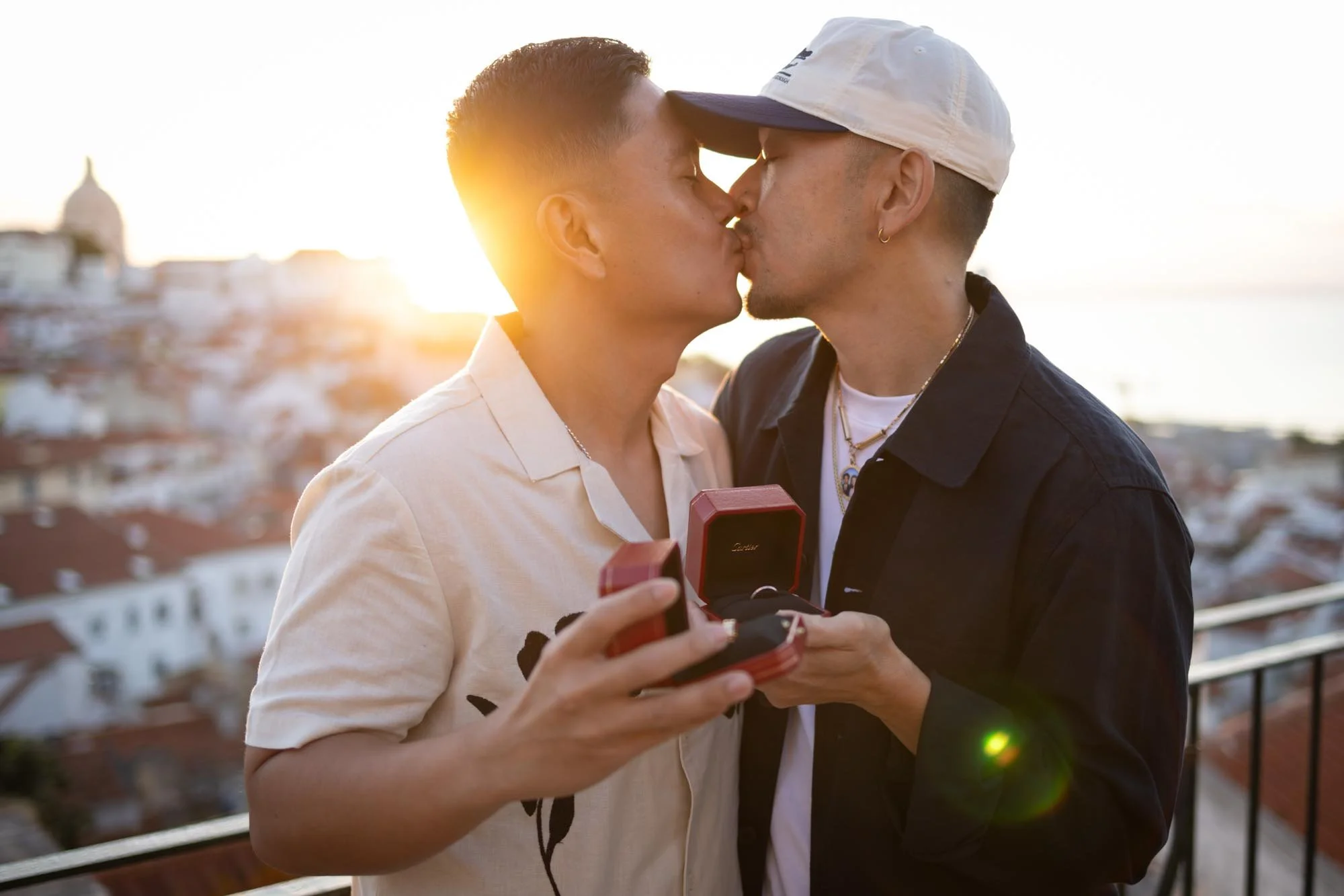 Two men kiss during sunset on a rooftop, one holding a small red box likely containing an engagement ring.