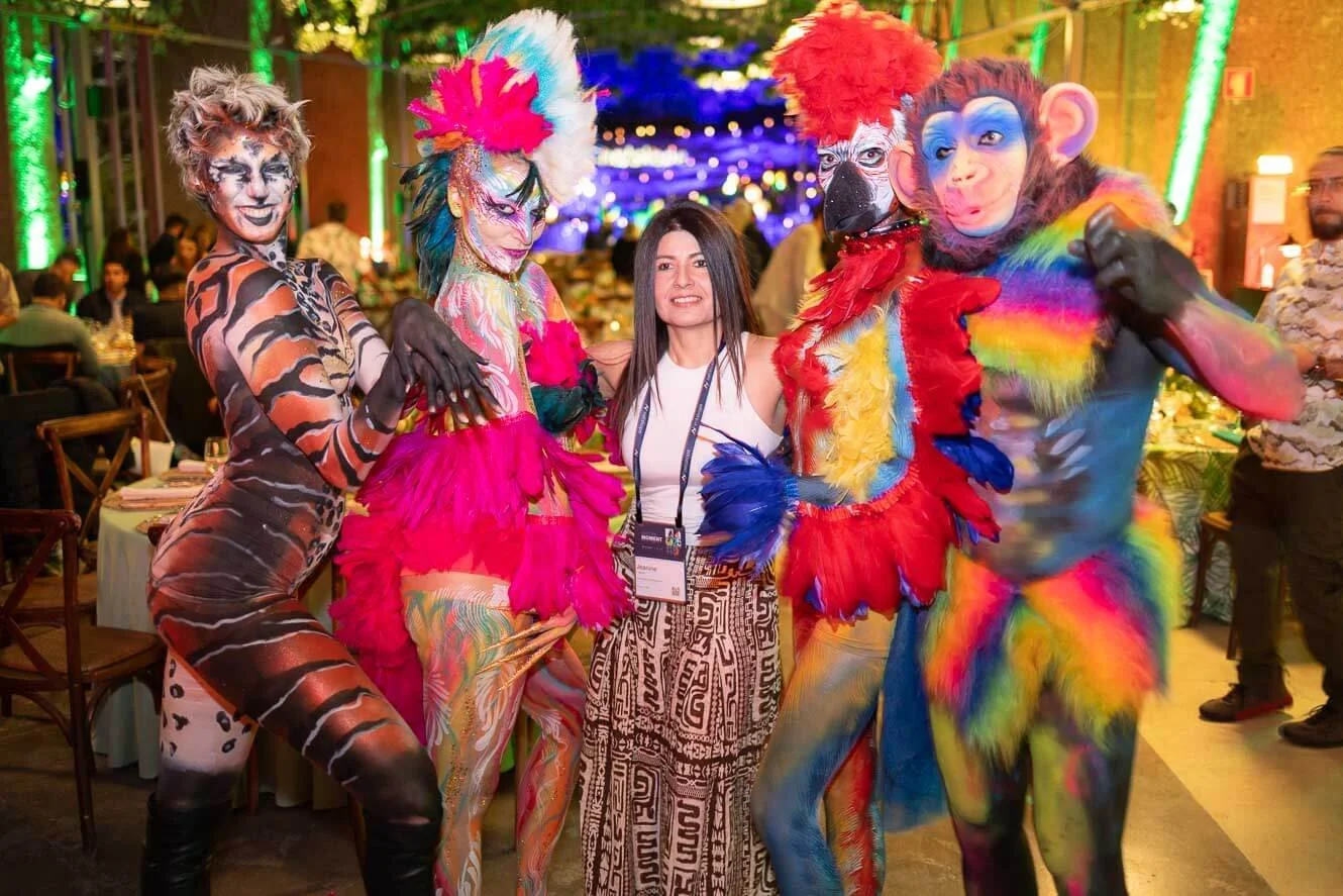 A woman in a white sleeveless top and patterned pants standing among four people dressed in colorful animal costumes with feathers and face paint, inside a decorated event hall with people and tables in the background.