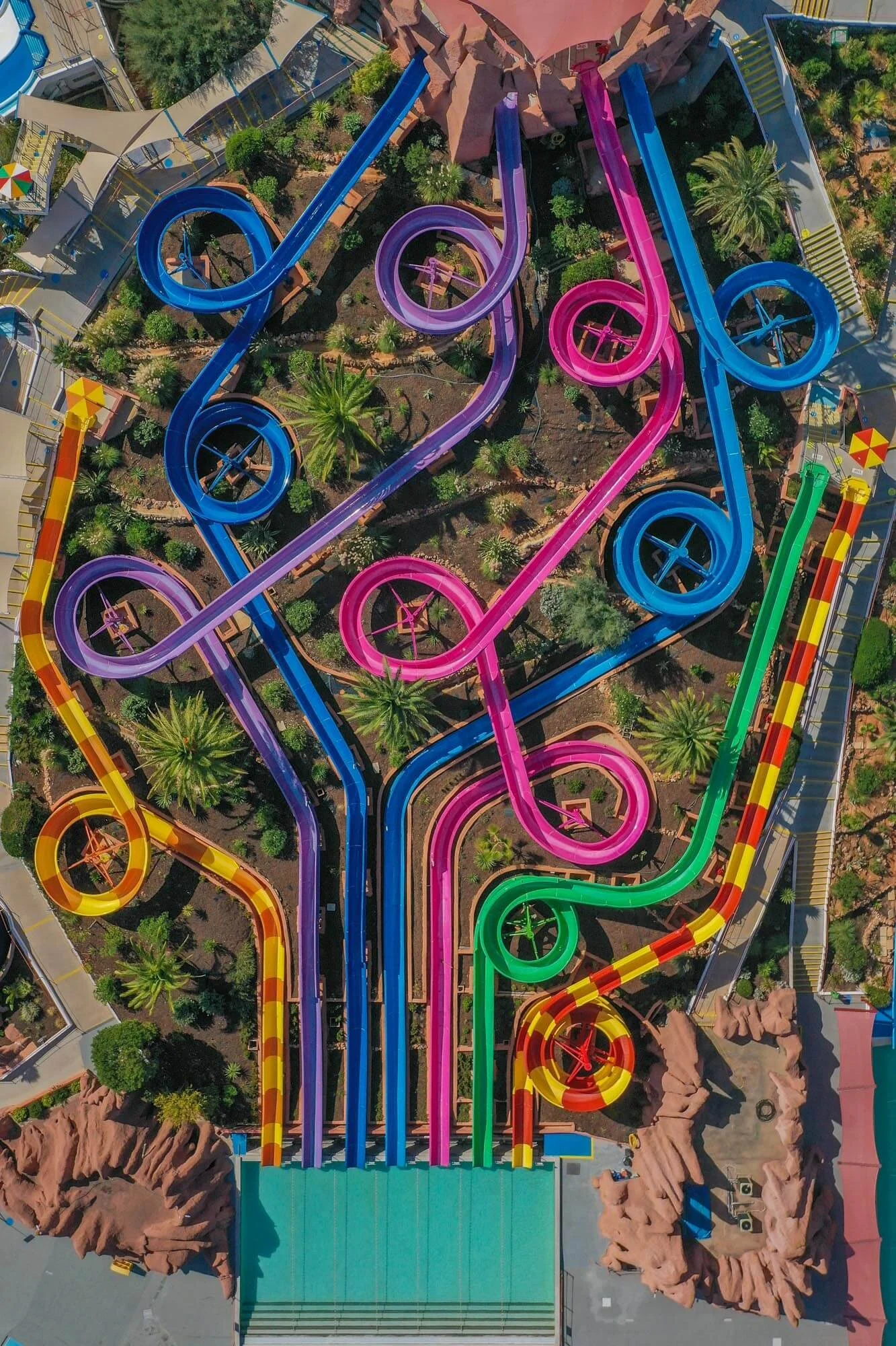 Aerial view of colorful water slides at a water park, with multiple intertwined slides in blue, pink, purple, yellow, and green, surrounded by trees and pathways.