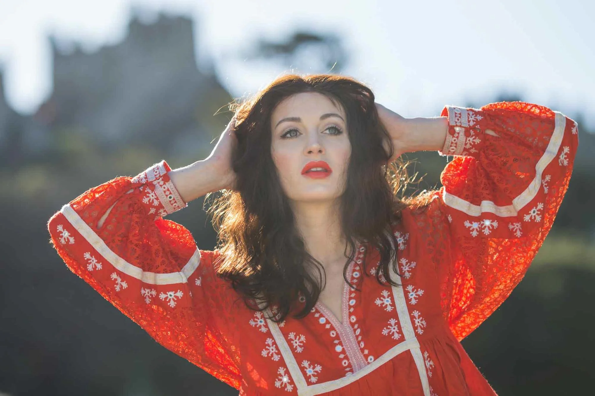 Woman with dark, wavy hair wearing a red embroidered dress, standing outdoors with her hands behind her head, looking to the side.