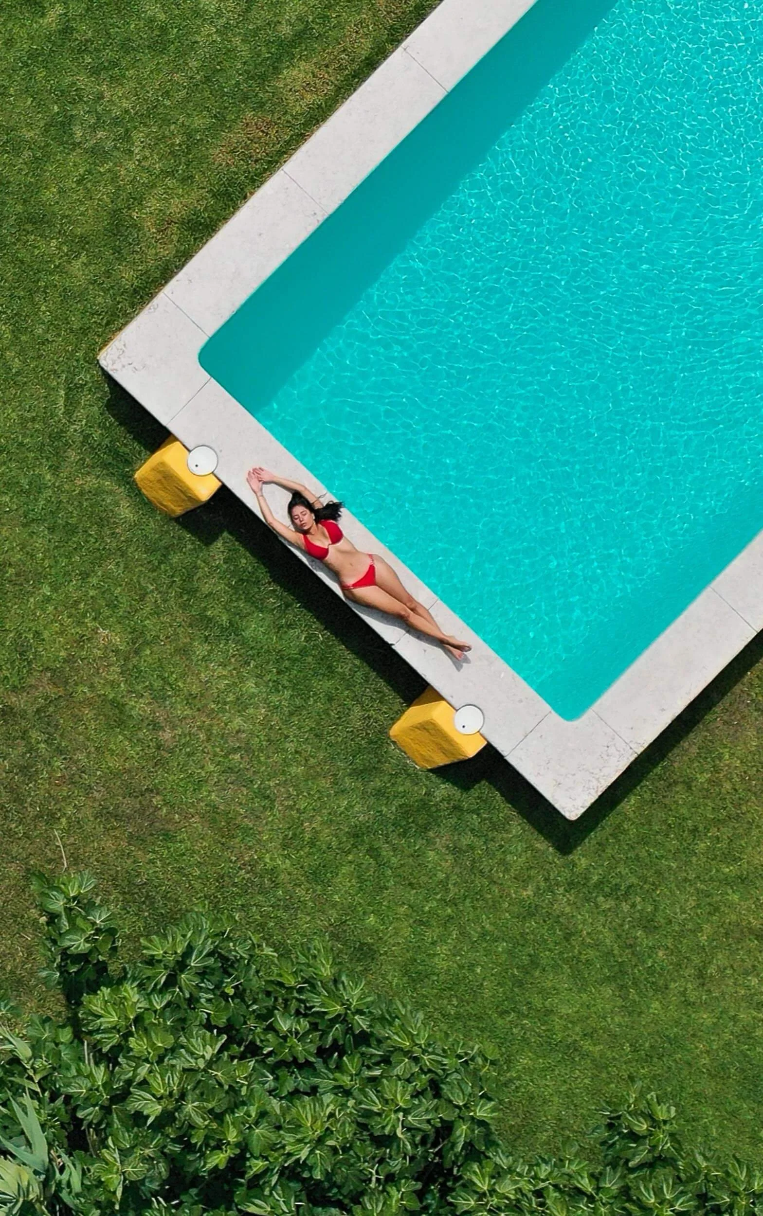 A woman in a red bikini lying on a poolside ledge with her arms above her head, next to a rectangular swimming pool surrounded by grass and some bushes.