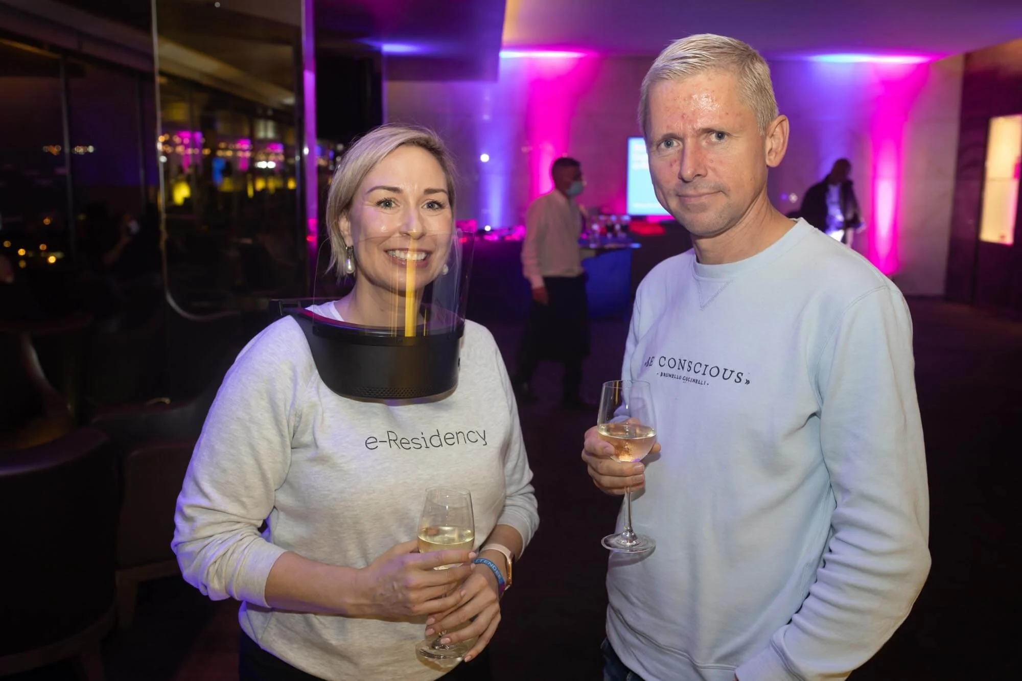 A woman and a man at a social event indoors, holding glasses of white wine, with colorful lighting, a large screen, and other people in the background.