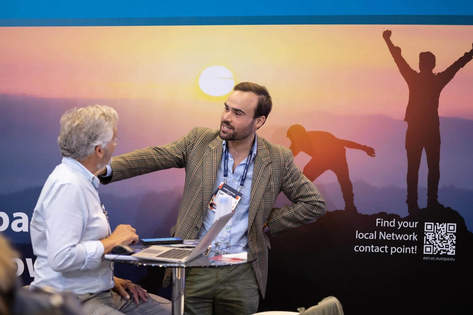 A man in a plaid blazer is engaging with an older man with glasses, who is sitting at a table with a laptop. Behind them, a large backdrop shows a sunrise or sunset with silhouettes of people on a mountain, one with arms raised and another bending ov