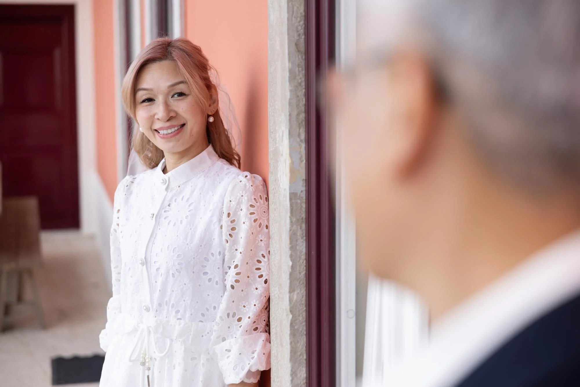 A woman with light skin and light brown hair in a white eyelet dress smiling and looking at another woman whose face is blurred in the foreground.