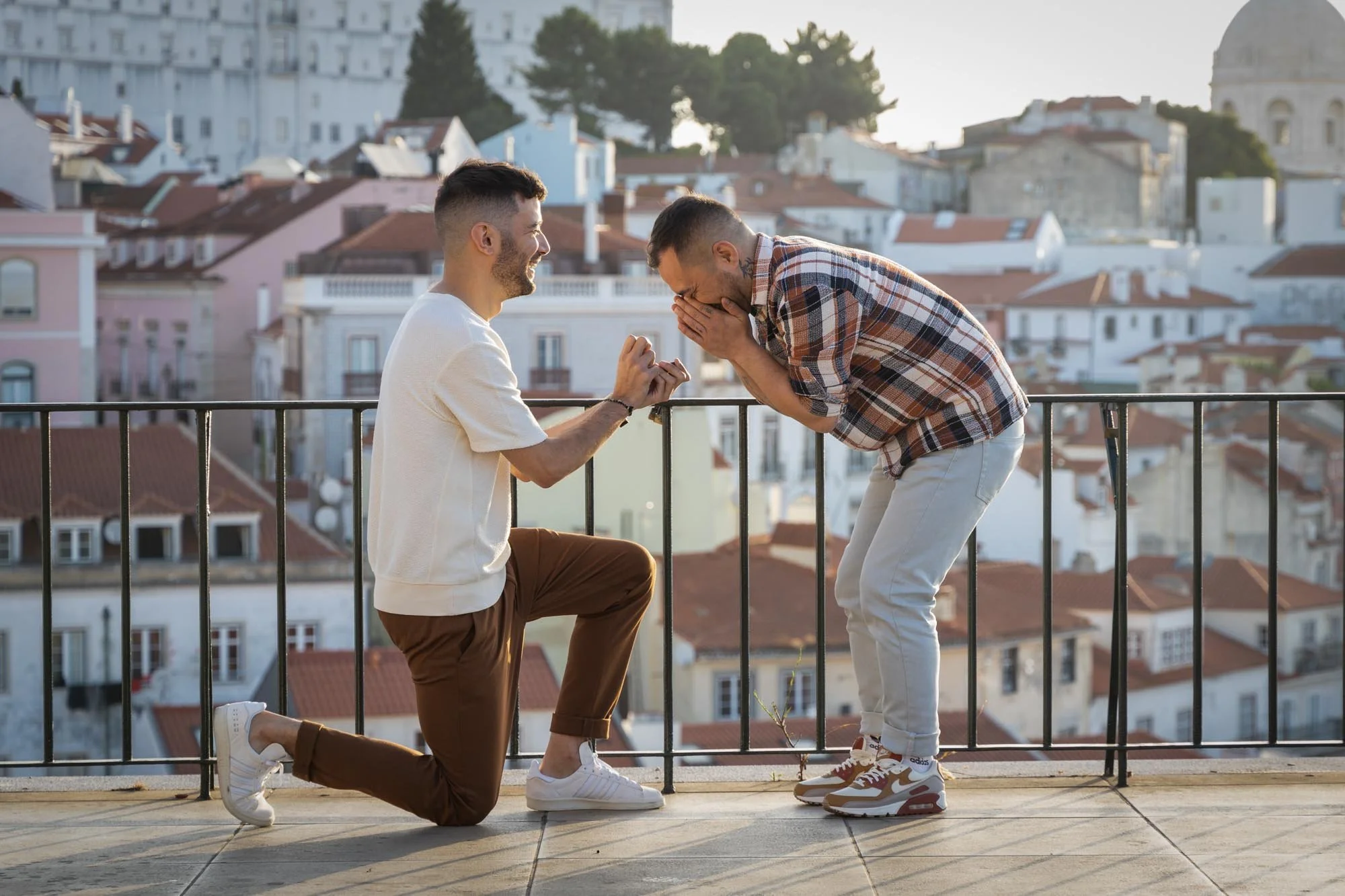 Two men are on a rooftop with a cityscape background. One man is kneeling on one knee, proposing, while the other man is standing and appears to be surprised and happy.