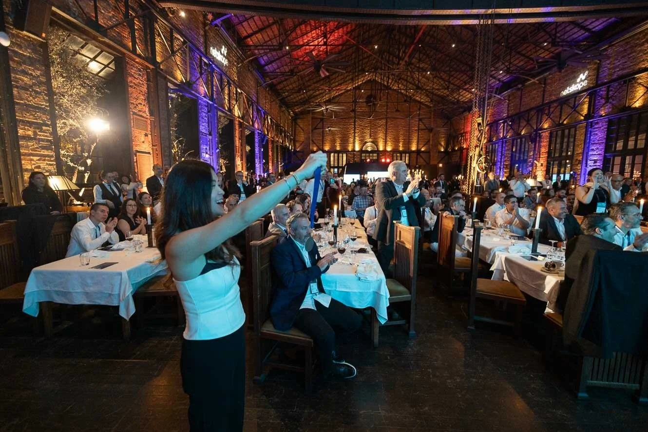 A large group of people in a dimly lit event hall with brick walls and high ceilings, attending a formal dinner or conference. Some people are standing, clapping, or taking pictures, while others are seated at tables with white tablecloths, glasses, 