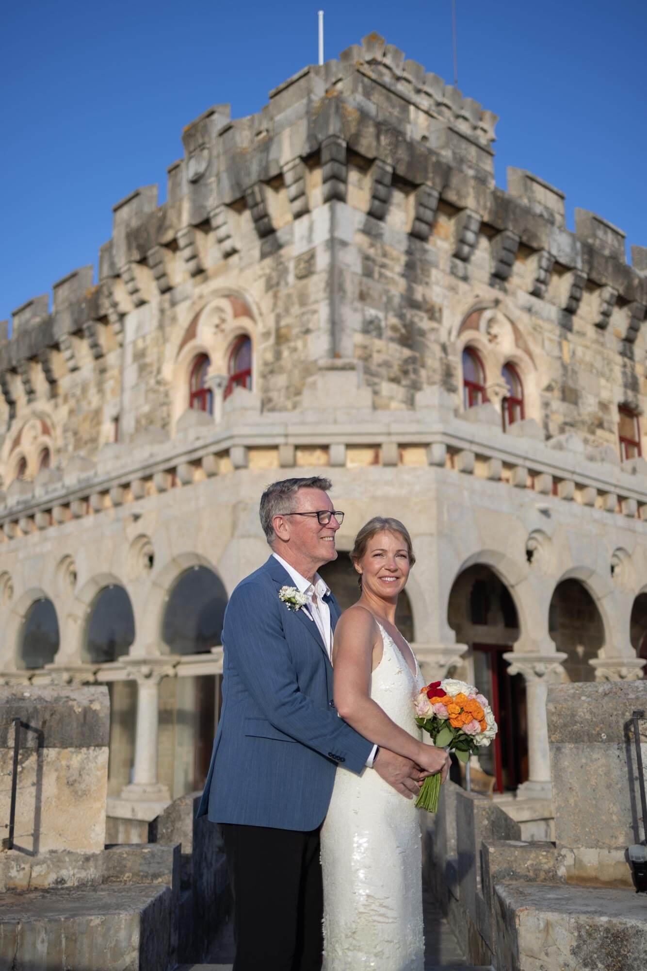 A couple dressed in wedding attire, standing outdoors in front of an old stone castle, holding a bouquet of flowers, during daytime.