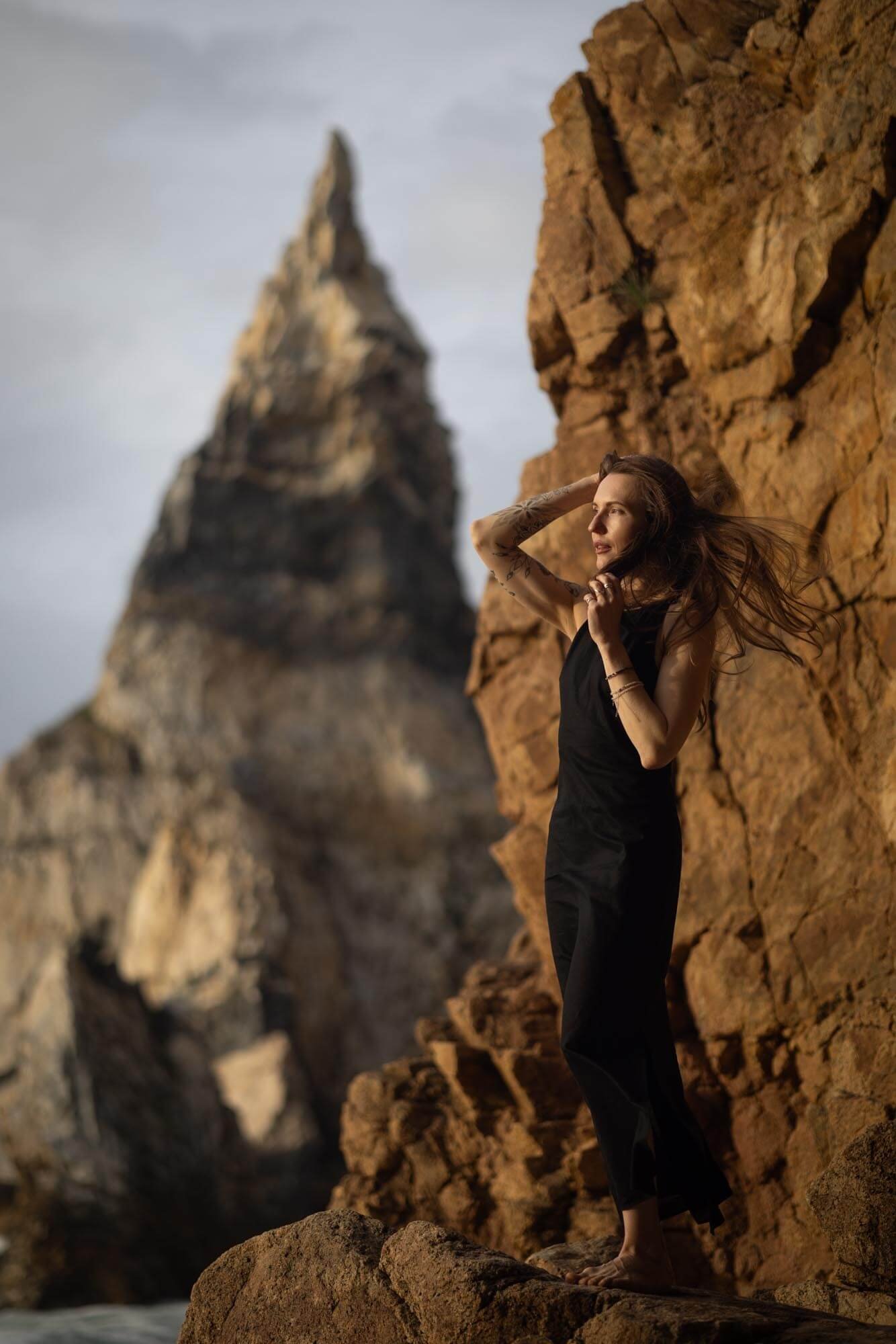 A woman with long hair and tattoos on her arms standing on rocks near a cliff, with a mountain summit in the background.