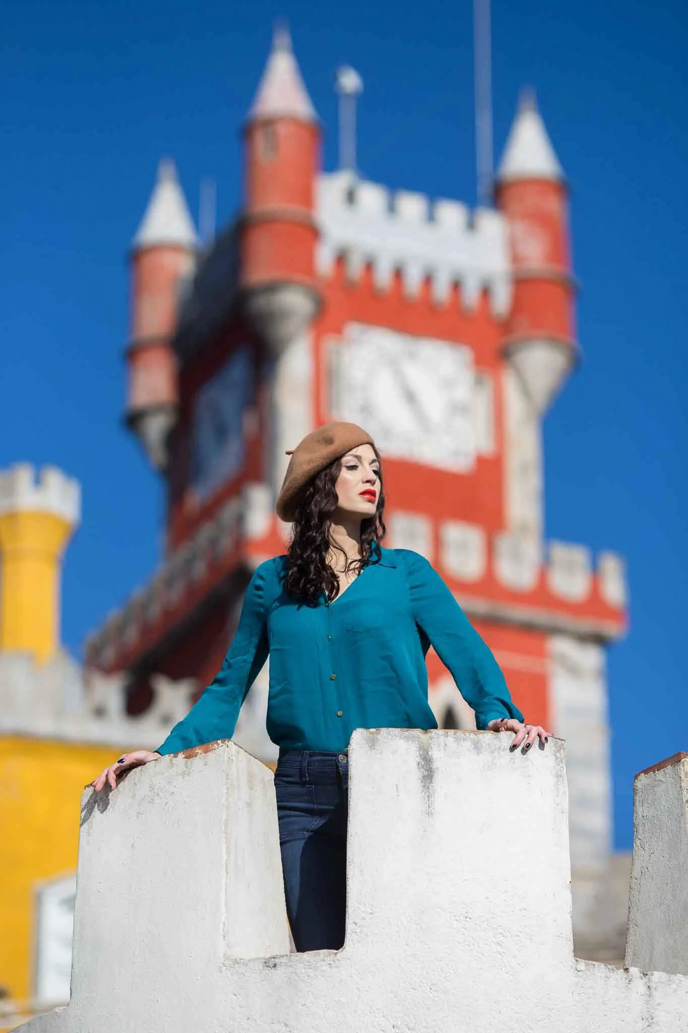 A woman with dark curly hair, wearing a tan beret and teal blouse, stands behind a white stone barrier with a colorful castle in the background on a bright day.