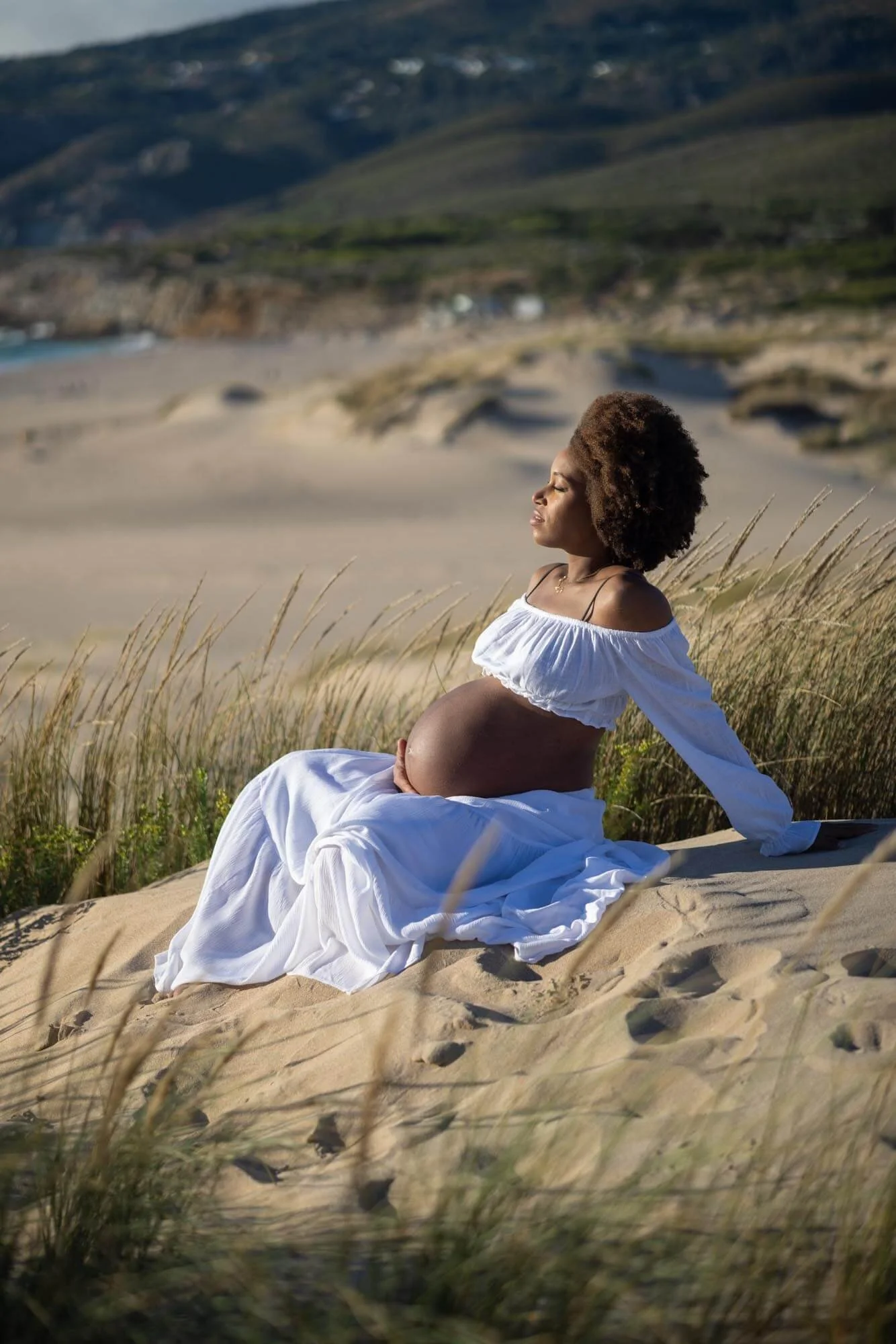 Pregnant woman sitting on sandy dunes at the beach, wearing a white off-shoulder dress, with her hand resting on her belly and eyes closed, surrounded by tall grass, with the ocean and hills in the background.
