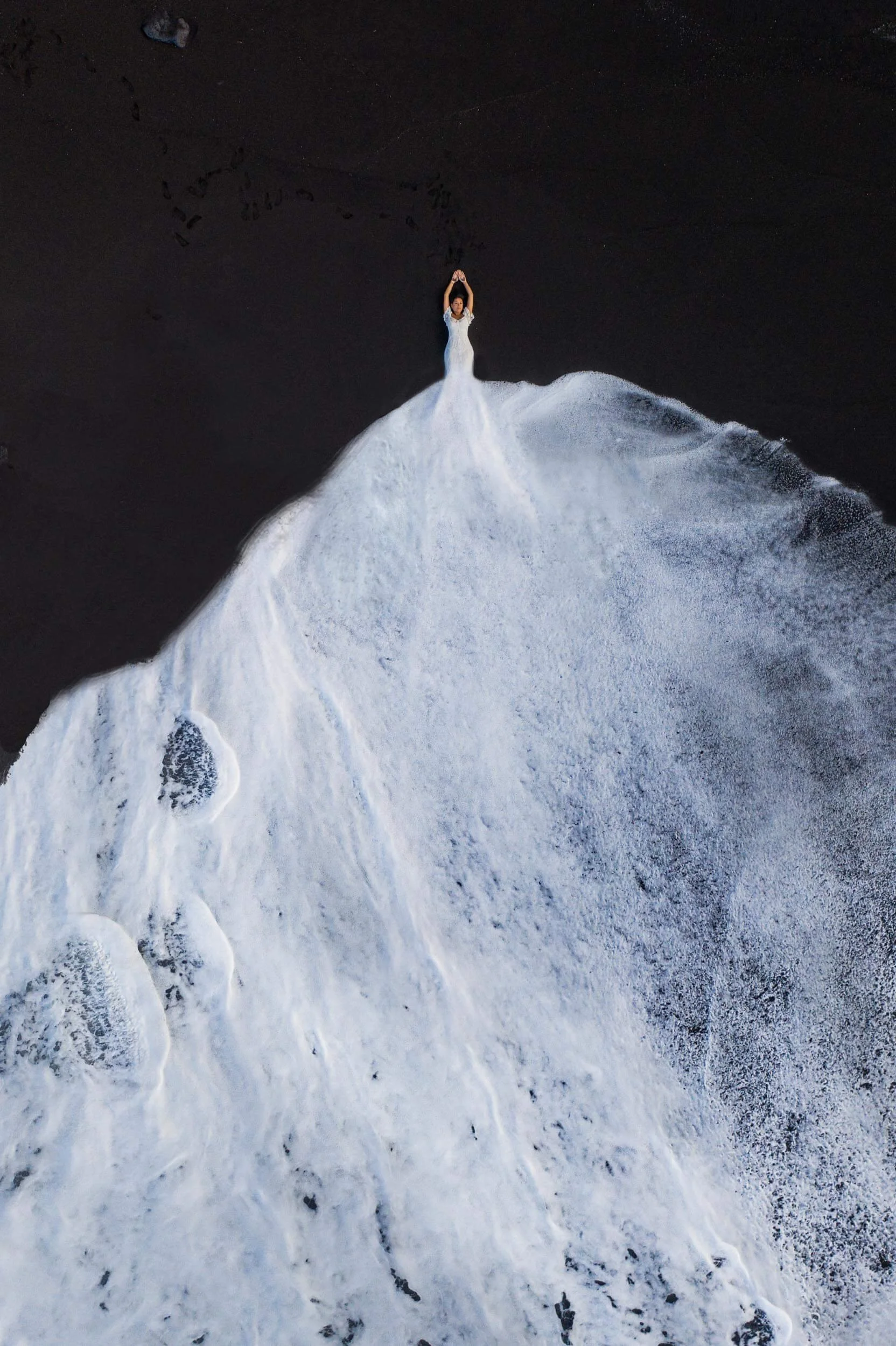 A woman in a white dress standing on a dark, wet sandy beach with waves washing over her, creating a visual illusion of her standing on a snow-covered mountain.