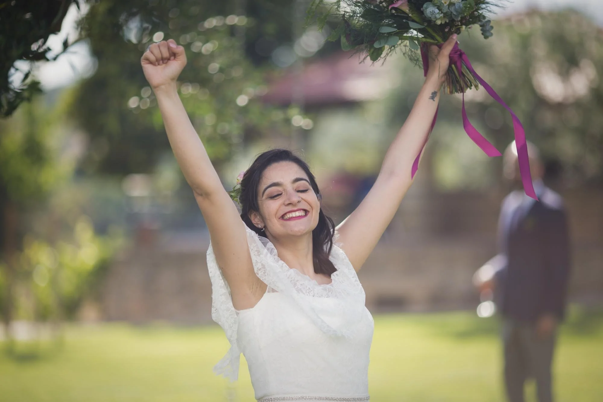 A woman in a white dress, smiling with eyes closed, raising her arms triumphantly holding a bouquet of flowers in an outdoor park setting, with a man in a suit standing in the background.