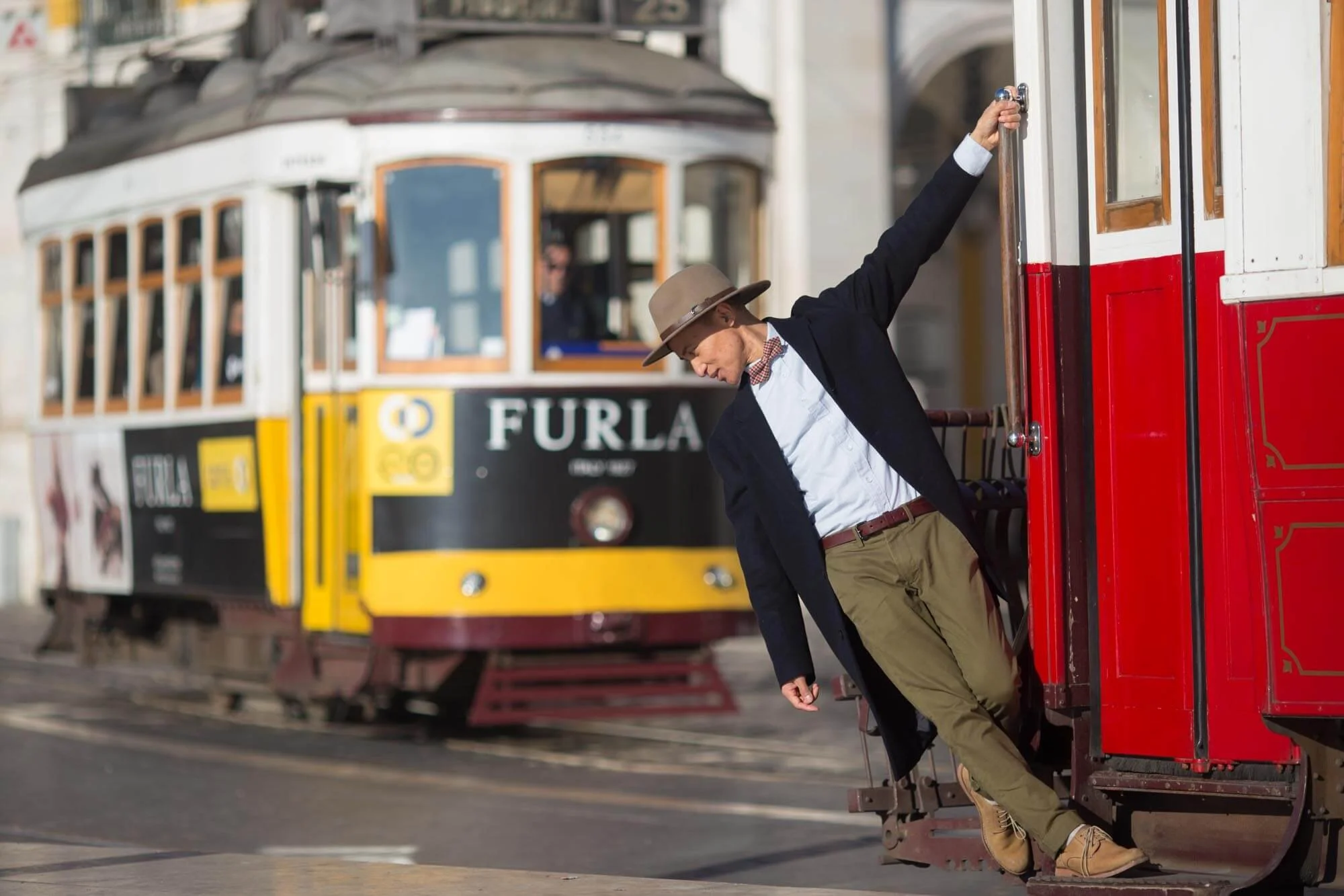 A man in vintage attire standing in the entrance of a red trolley car, with a classic yellow and black tram in the background.