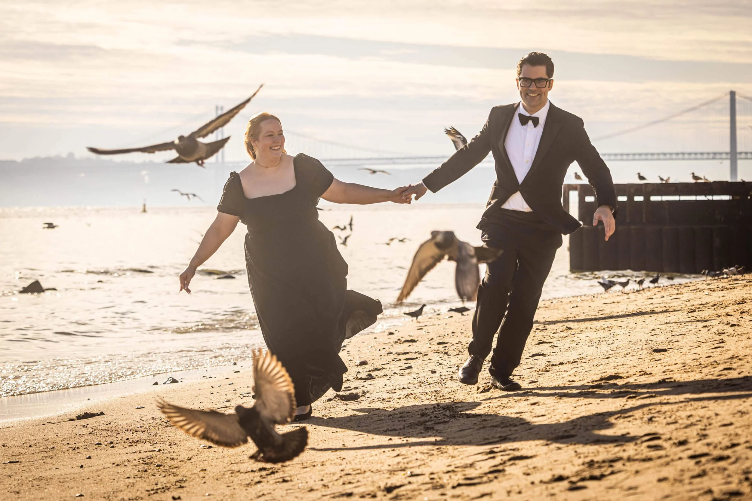 Lisbon engagement photoshoot with a happy couple running between the birds in a formal outfit with the bridge in the the background.