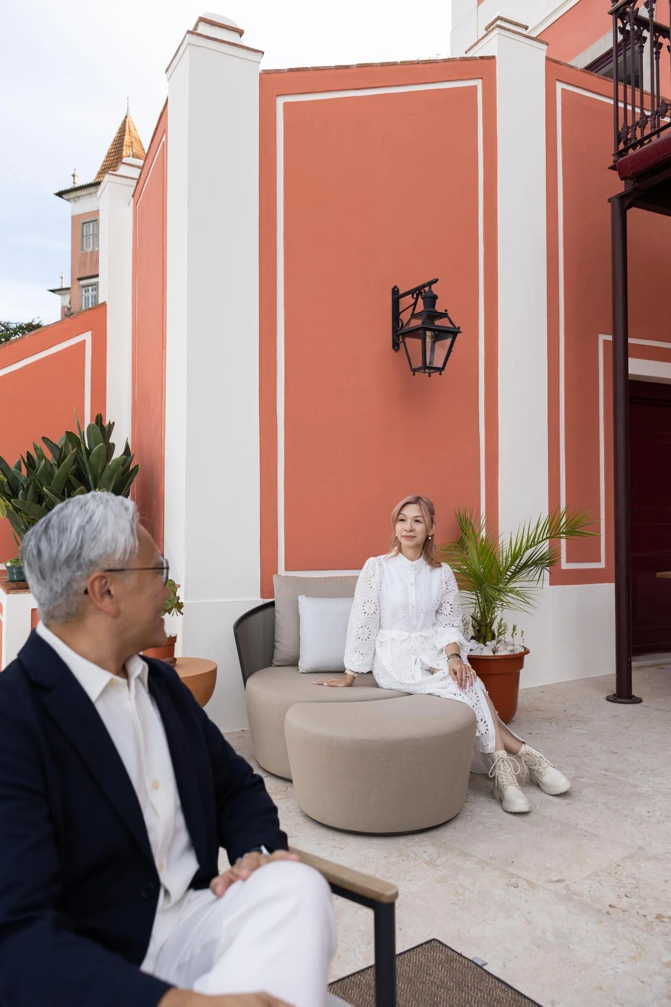 A woman in a white dress seated on a modern beige outdoor sofa with white cushions, next to potted plants, and a man with gray hair and glasses sitting nearby outdoors against a colorful pink and white wall with a lantern-style light fixture.