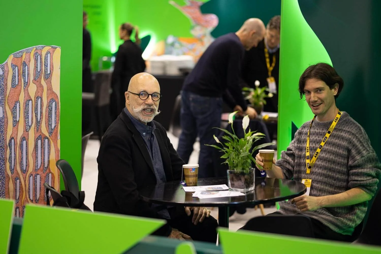 Two men sitting at a round black table with a small green plant in a pot, holding coffee cups, in a vibrant green interior space at a conference or event. One man has a bald head, glasses, and a beard, dressed in a dark jacket. The other man has dark