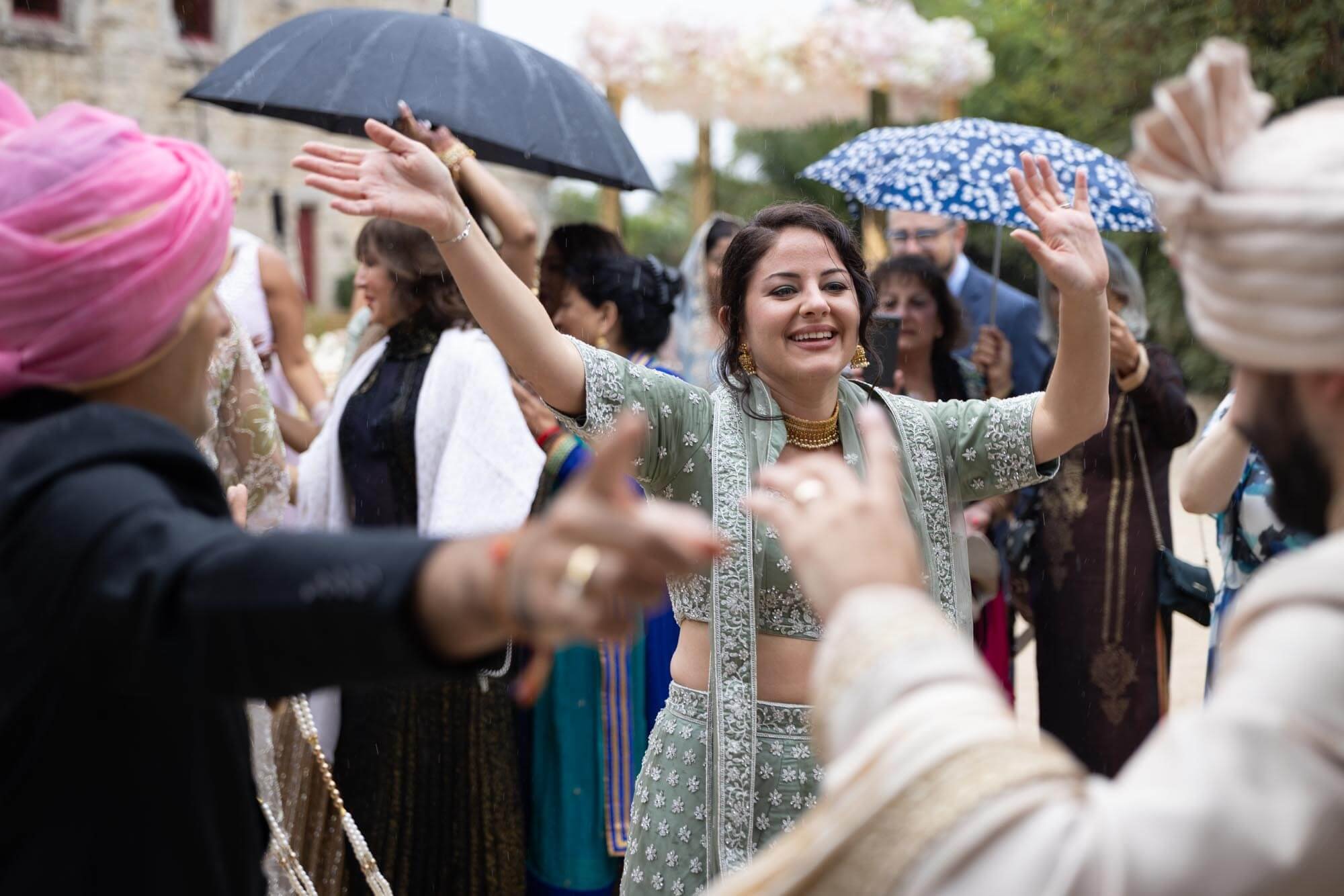 People celebrating at an outdoor event, some wearing traditional South Asian attire, with umbrellas and smiles.