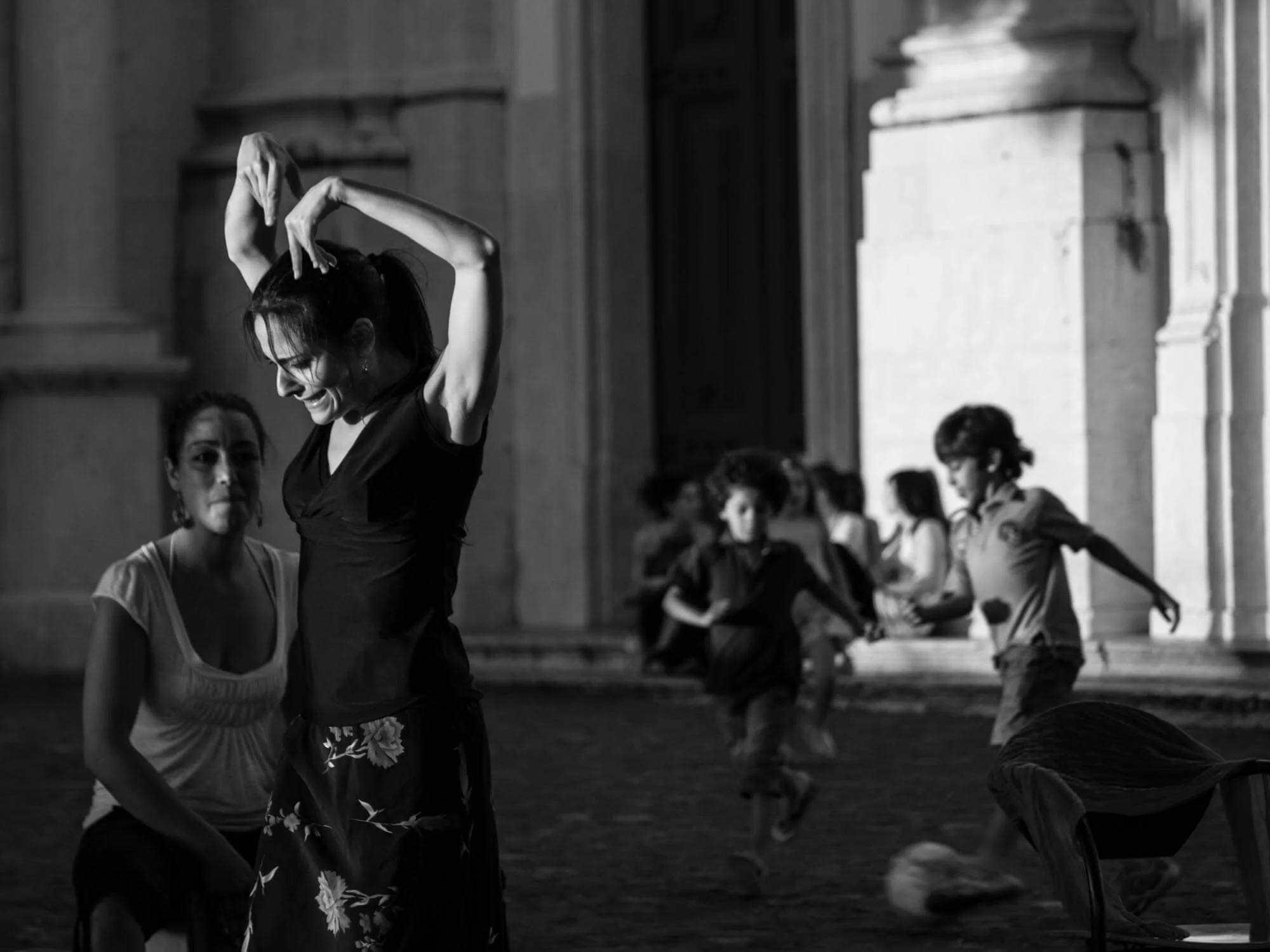 Women dancing indoors in a black and white photo, with one woman in the foreground smiling and posing with arms raised, and several women in the background engaged in dance or movement.