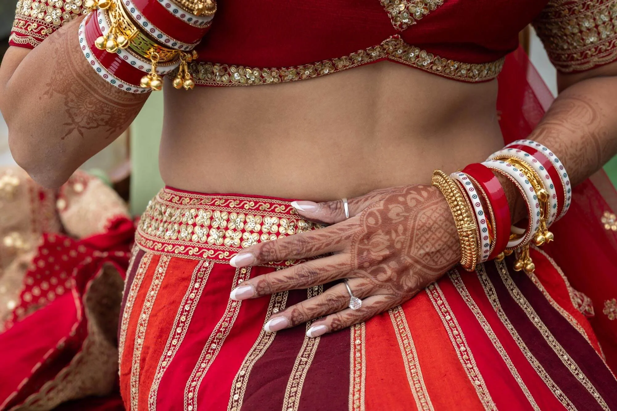 Close-up of a woman dressed in traditional Indian attire, showing her midriff, adorned with jewelry, henna on her hands, and a red and gold embroidered skirt.