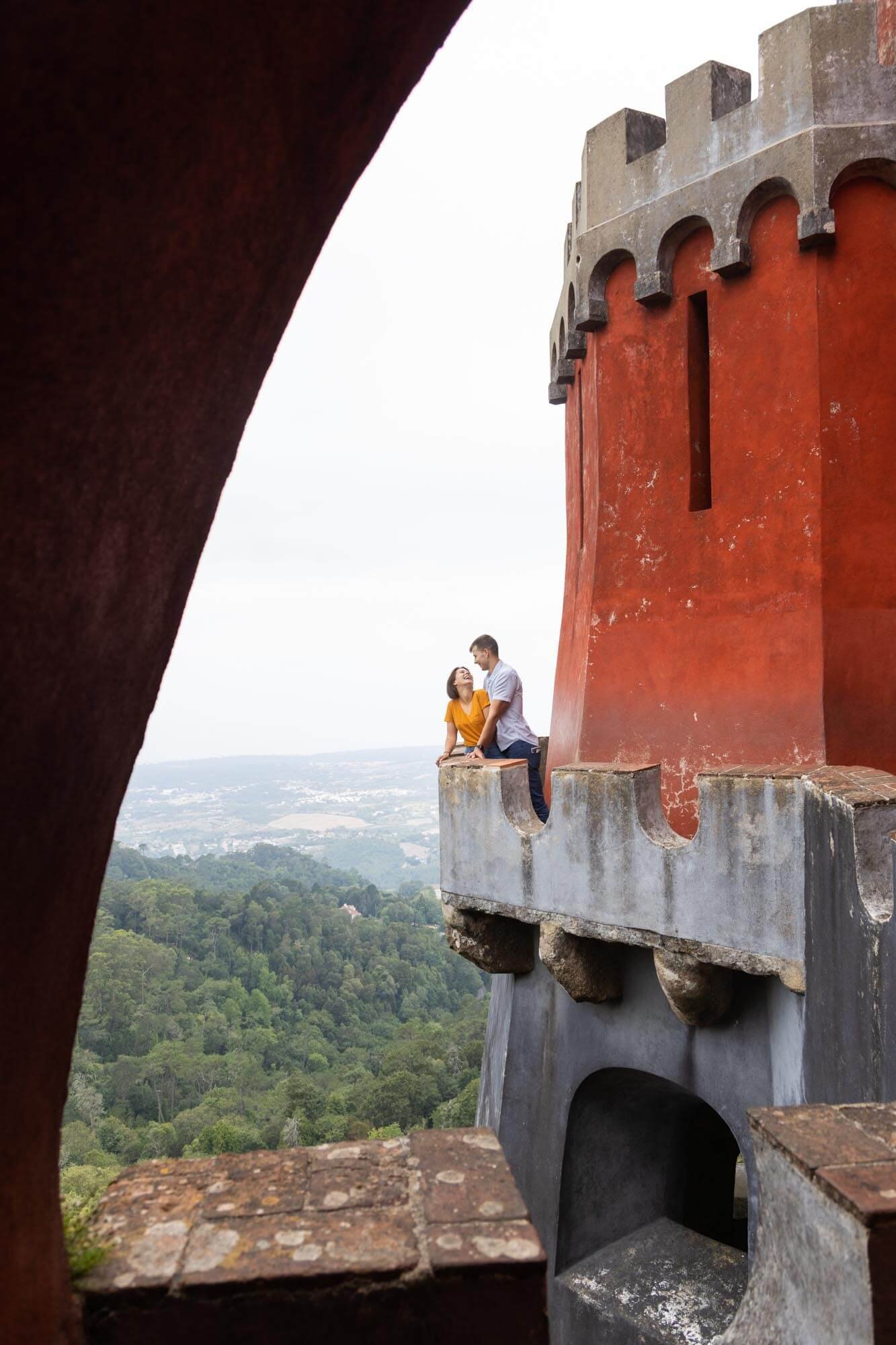 A couple stands on a castle tower balcony overlooking a forested landscape with distant hills.