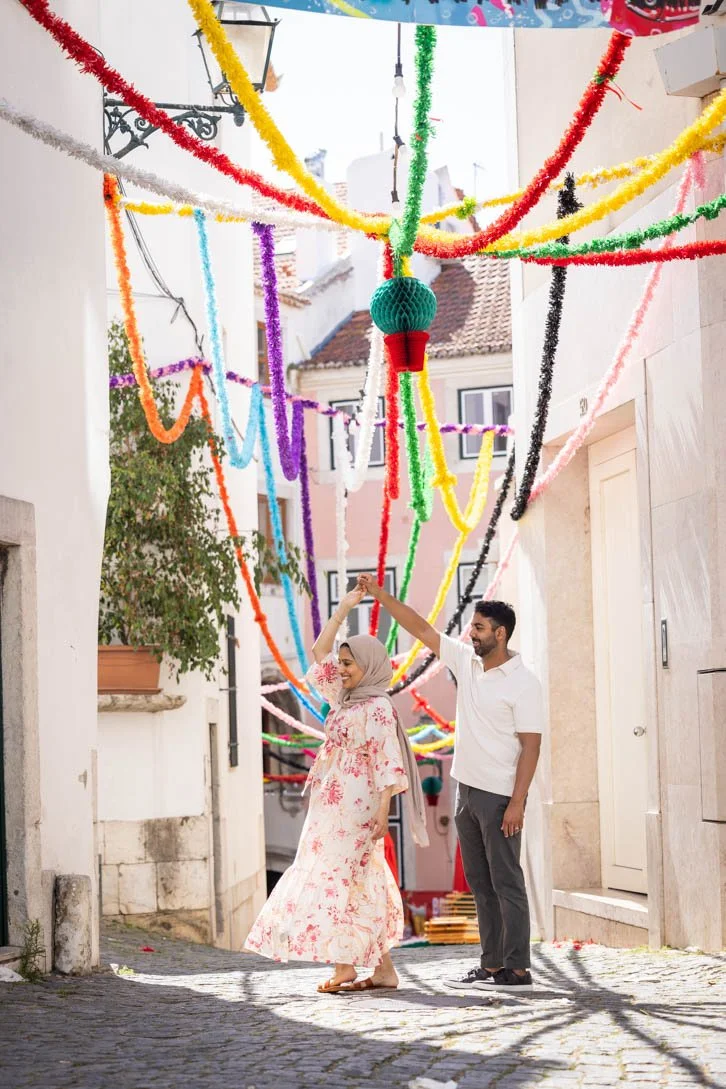A couple is posing for a photoshoot, dancing under colorful streamers hanging above on a narrow street of Alfama