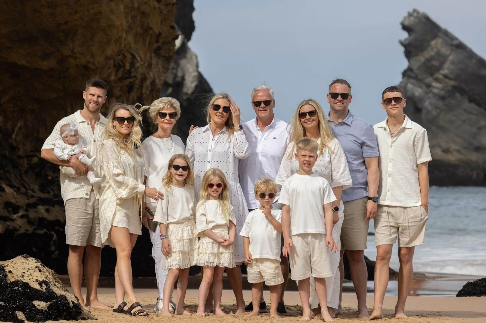 Family group photo on a beach with rocky cliffs in the background, all dressed in light-colored clothing.