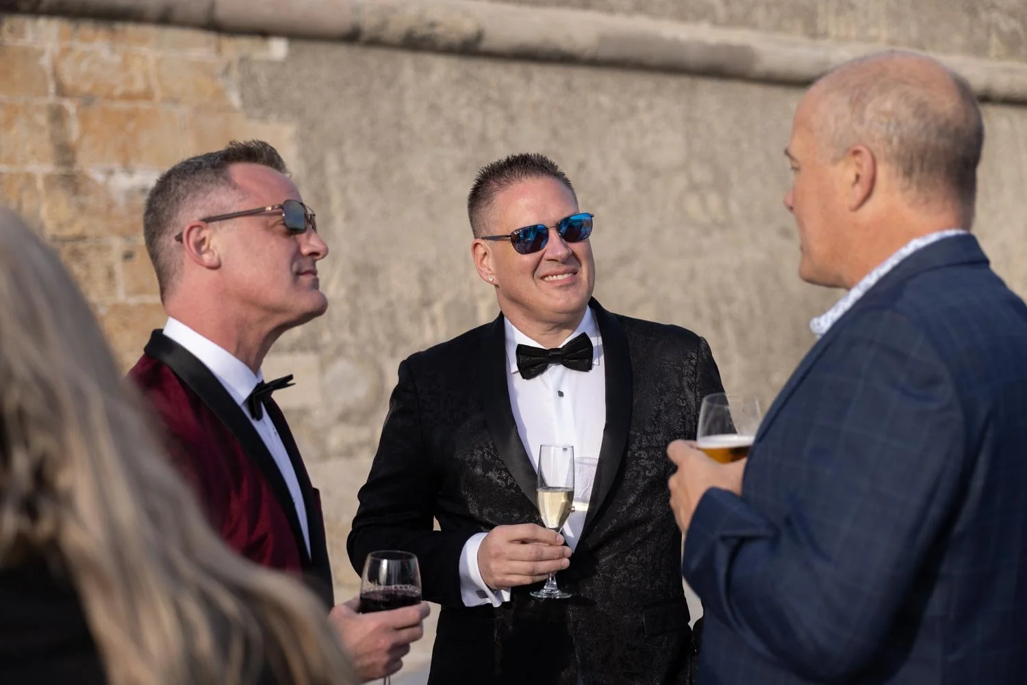 Three men in tuxedos and sunglasses are having a conversation at a formal event, holding wine glasses, with a brick wall in the background.