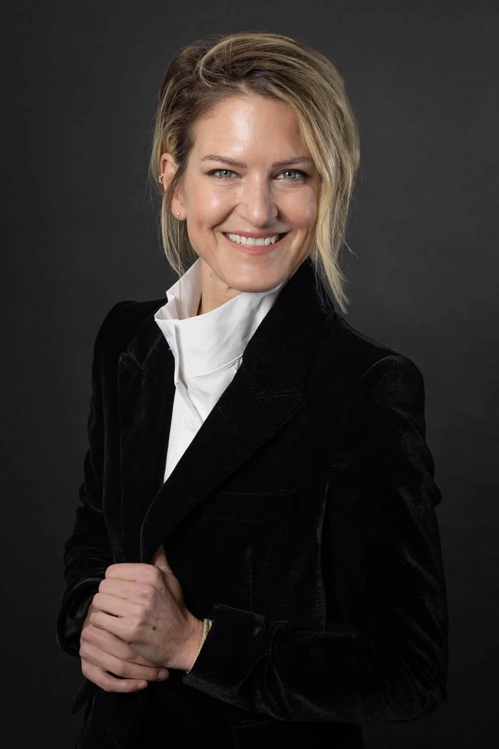 Portrait of a woman with blonde hair in a black blazer and white shirt smiling against a dark background.
