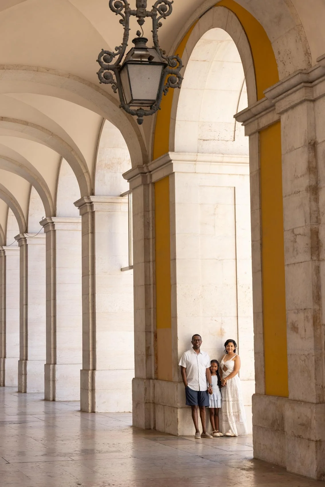 A family of three standing under a large archway in a historic building with beige stone walls and a decorative black lantern hanging from the ceiling.