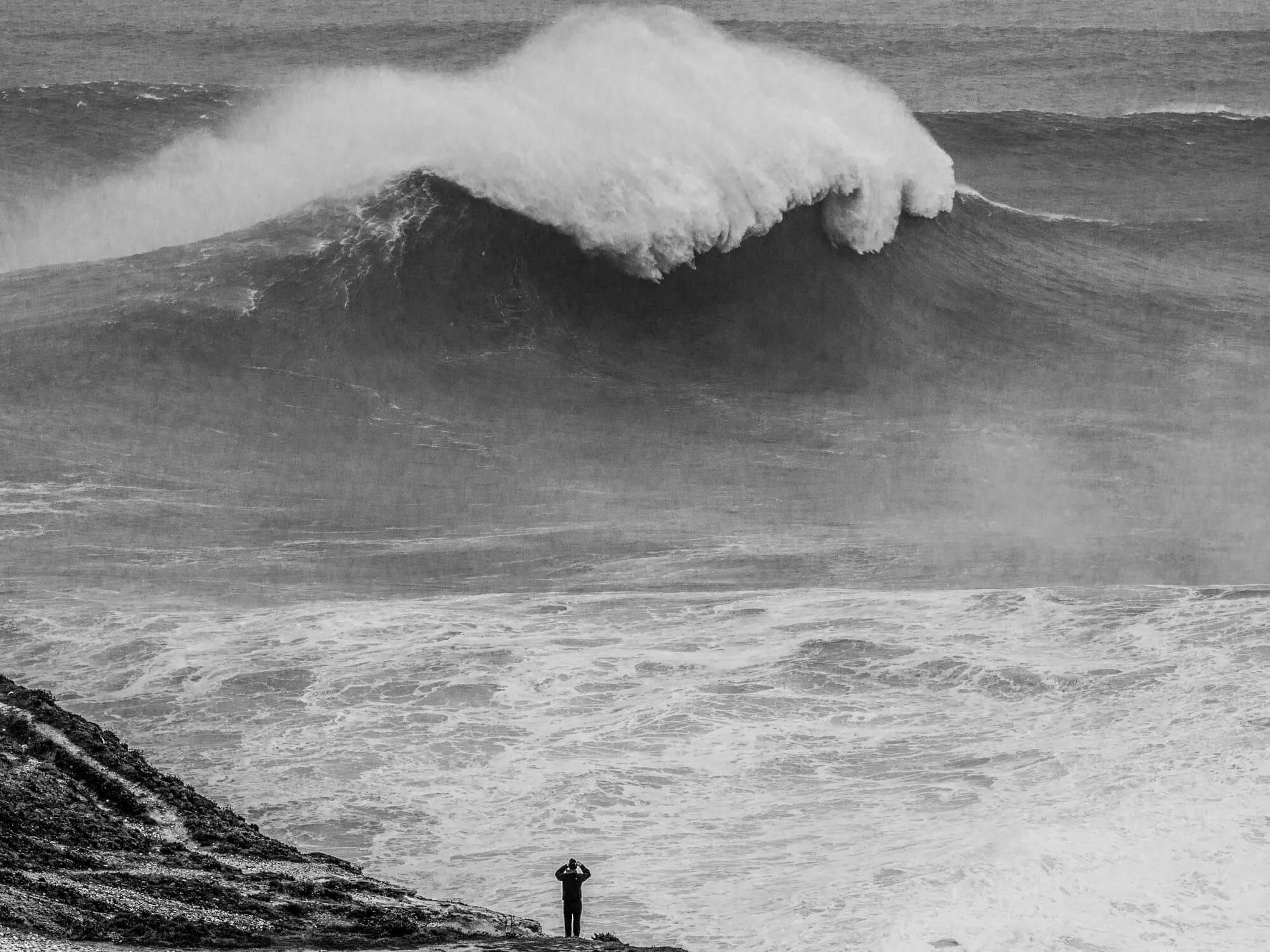 A person standing on a rocky shoreline taking a photograph of a large, powerful ocean wave in black and white.