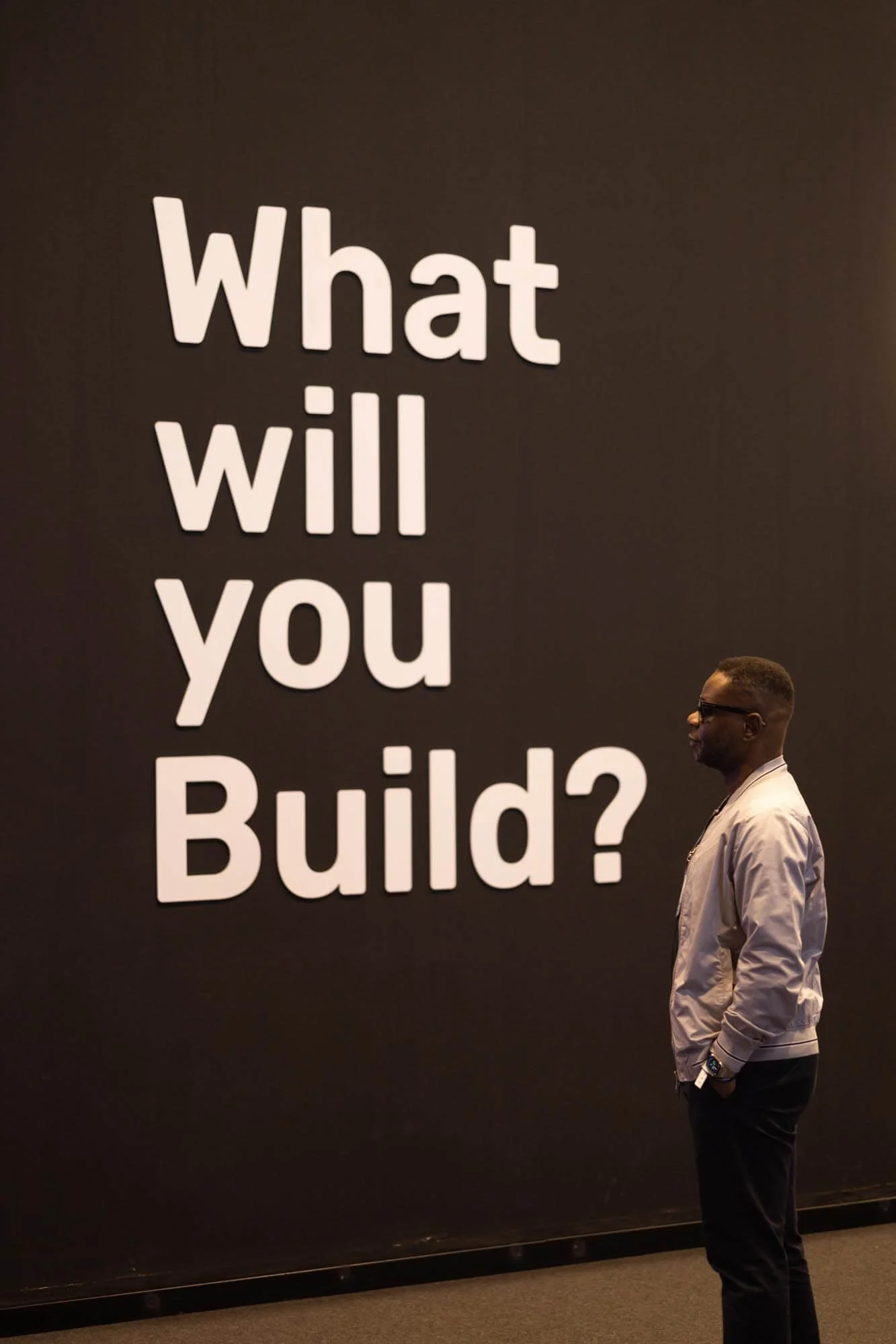 A man standing in profile looking at a large black wall with the white text 'What will you build?' on it.