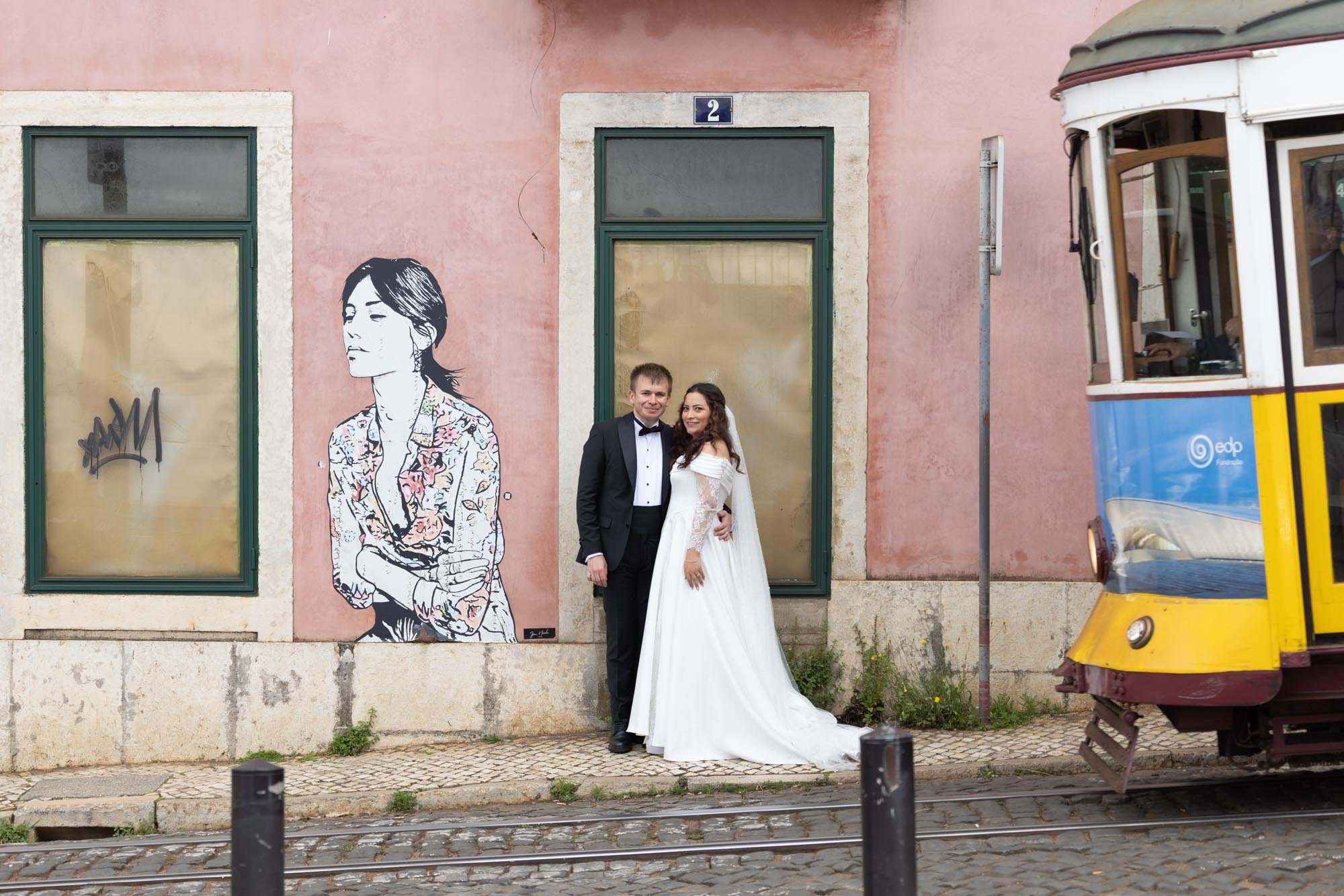 A happy couple in wedding attire standing on a cobblestone sidewalk in front of a pink building with green-framed windows and a mural of a woman on the wall. The groom wears a black tuxedo and the bride wears a white wedding dress. A yellow and blue 