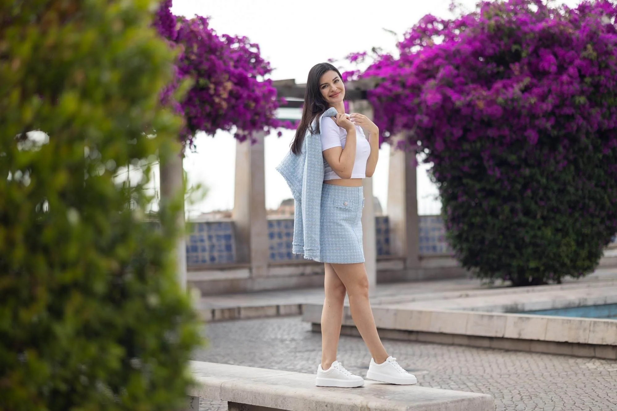 Young woman standing outdoors near purple flowering bushes, wearing a white crop top, light blue skirt, and white sneakers, smiling and carrying a light blue jacket over her shoulder.