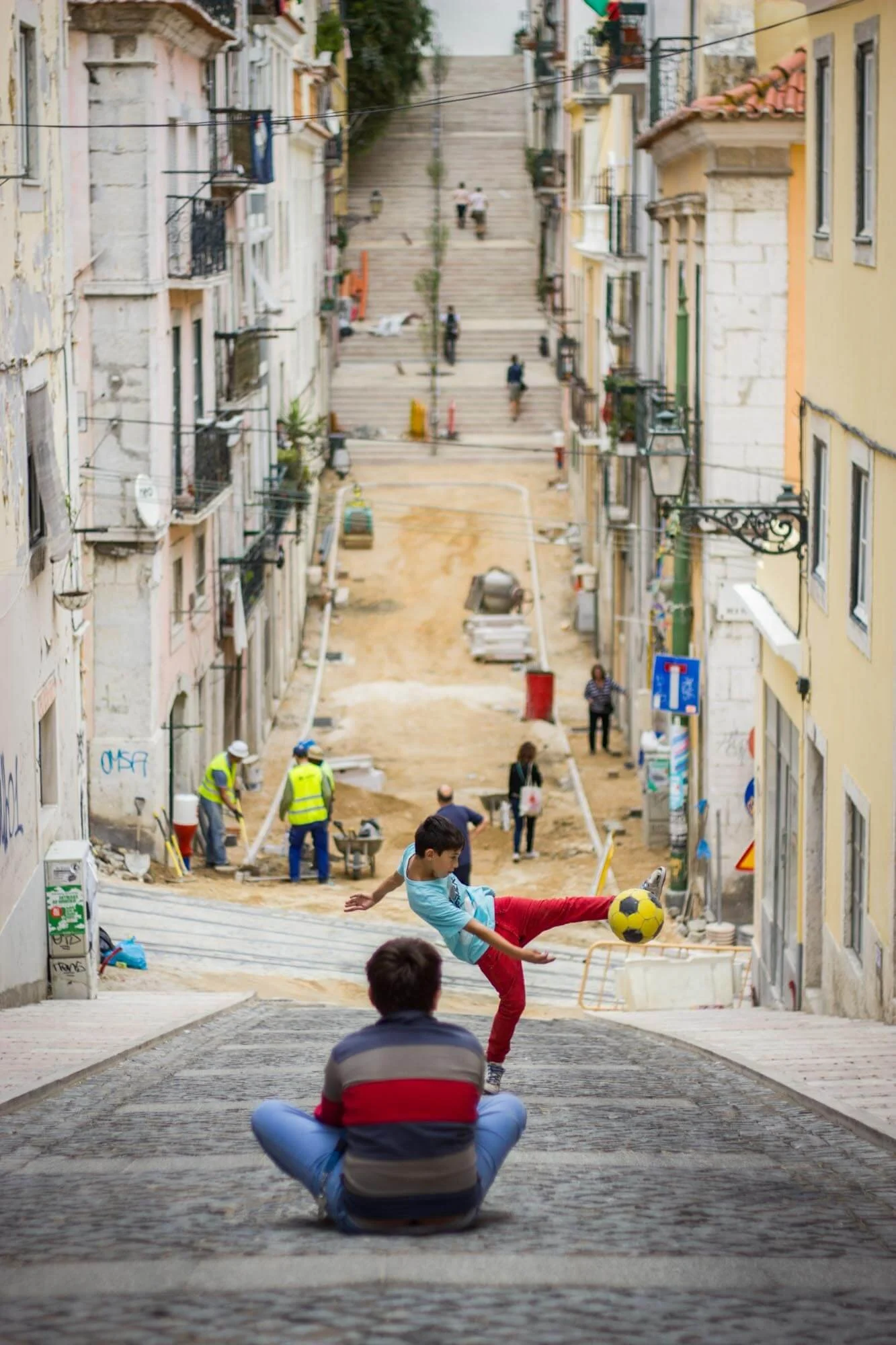 A boy playing soccer on a street with a man sitting on the ground watching. The street is under renovation with construction workers and equipment, and is lined with colorful buildings and steps leading uphill in the background.
