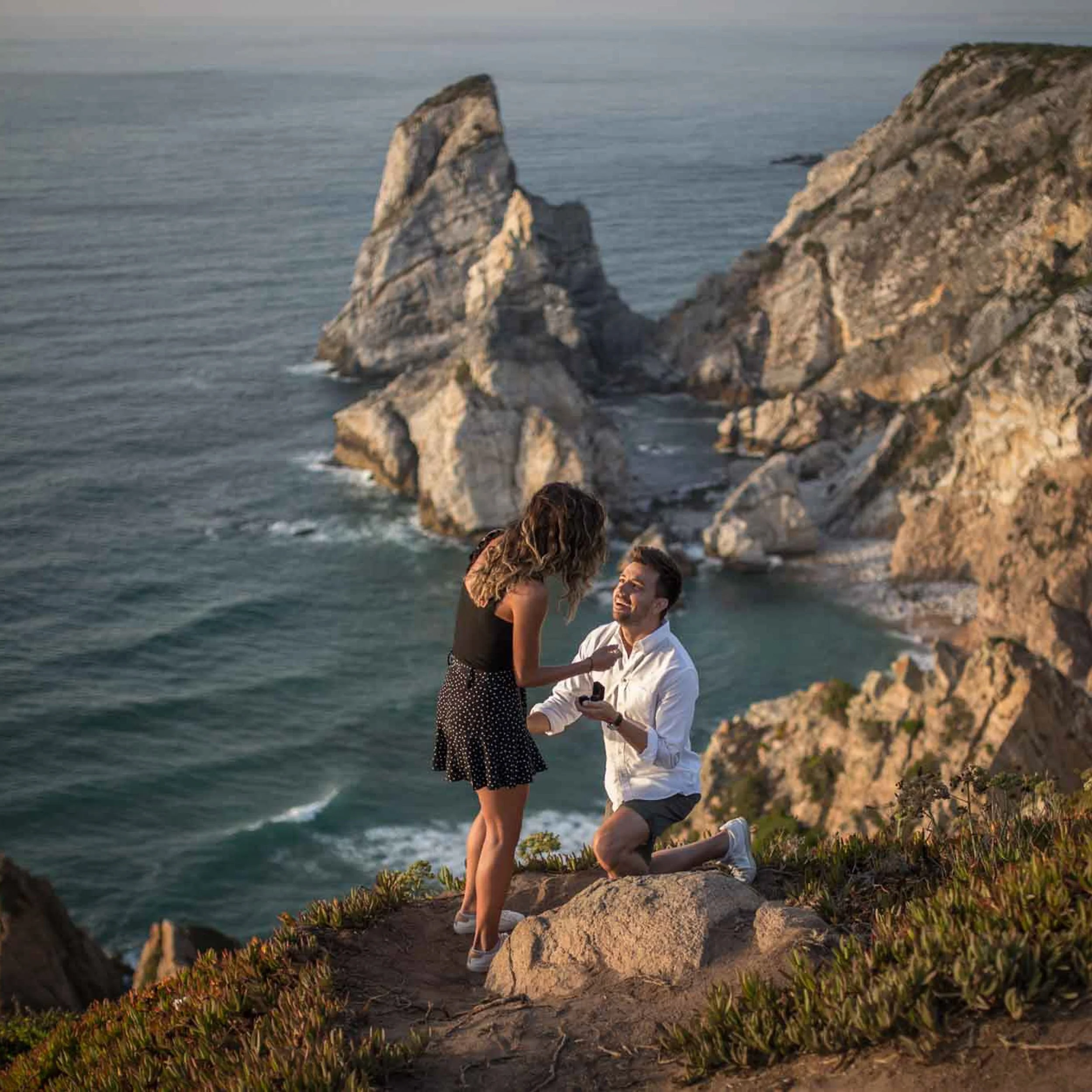 A man on one knee proposing marriage to a woman on a rocky cliff overlooking the Ursa beach, with rocky sea stacks in the background at sunset.