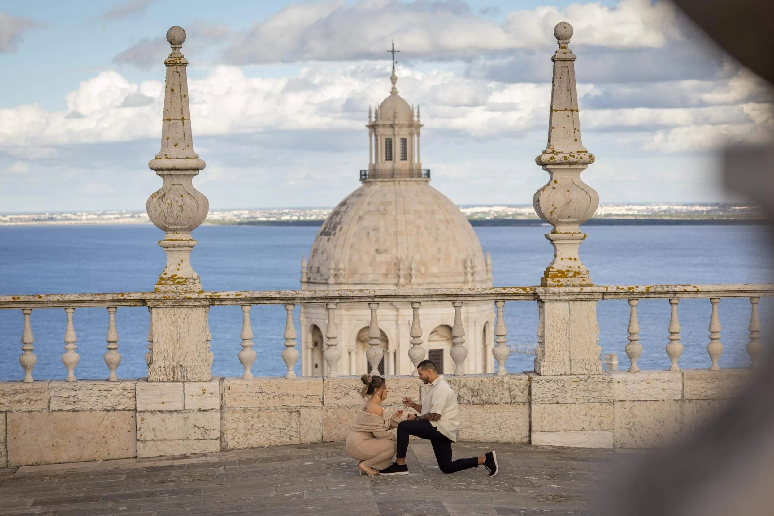 A man proposing marriage to a woman on a balcony overlooking a large domed building and water in the background.