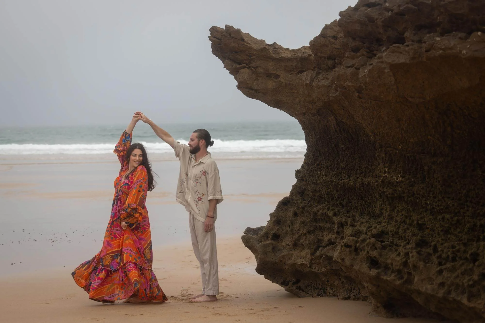 A man and woman dancing on a beach near a large rock formation, with the ocean in the background.