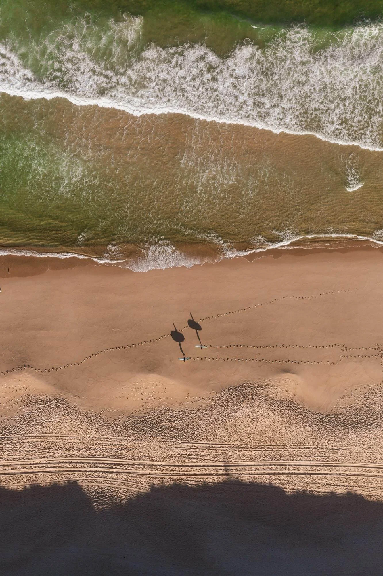 An aerial view of a beach with two umbrellas, towel, and footprints in the sand, with ocean waves crashing onto the shore.