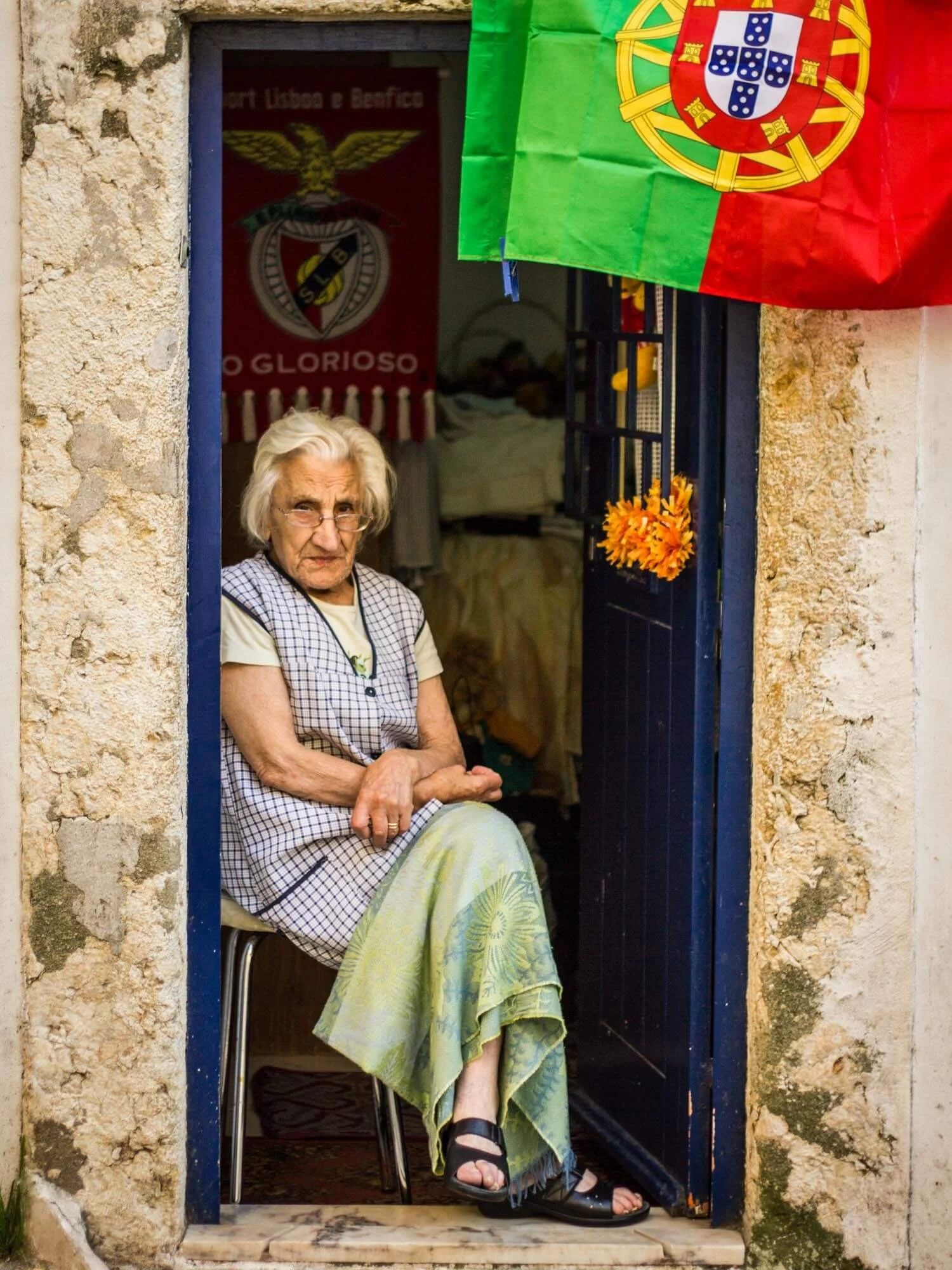 An elderly woman sitting in a doorway wearing glasses, a checkered dress, and a floral skirt, with Portuguese flags hanging outside.