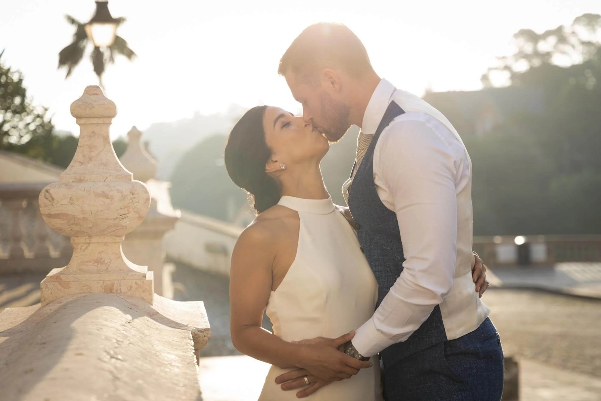 A couple kissing outdoors in warm sunlight, with a decorative stone railing and trees in the background.
