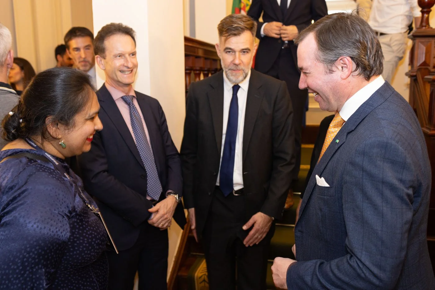 Group of five people, four men and one woman, engaged in conversation and smiling in a formal setting with wooden stairs and a flag in the background.
