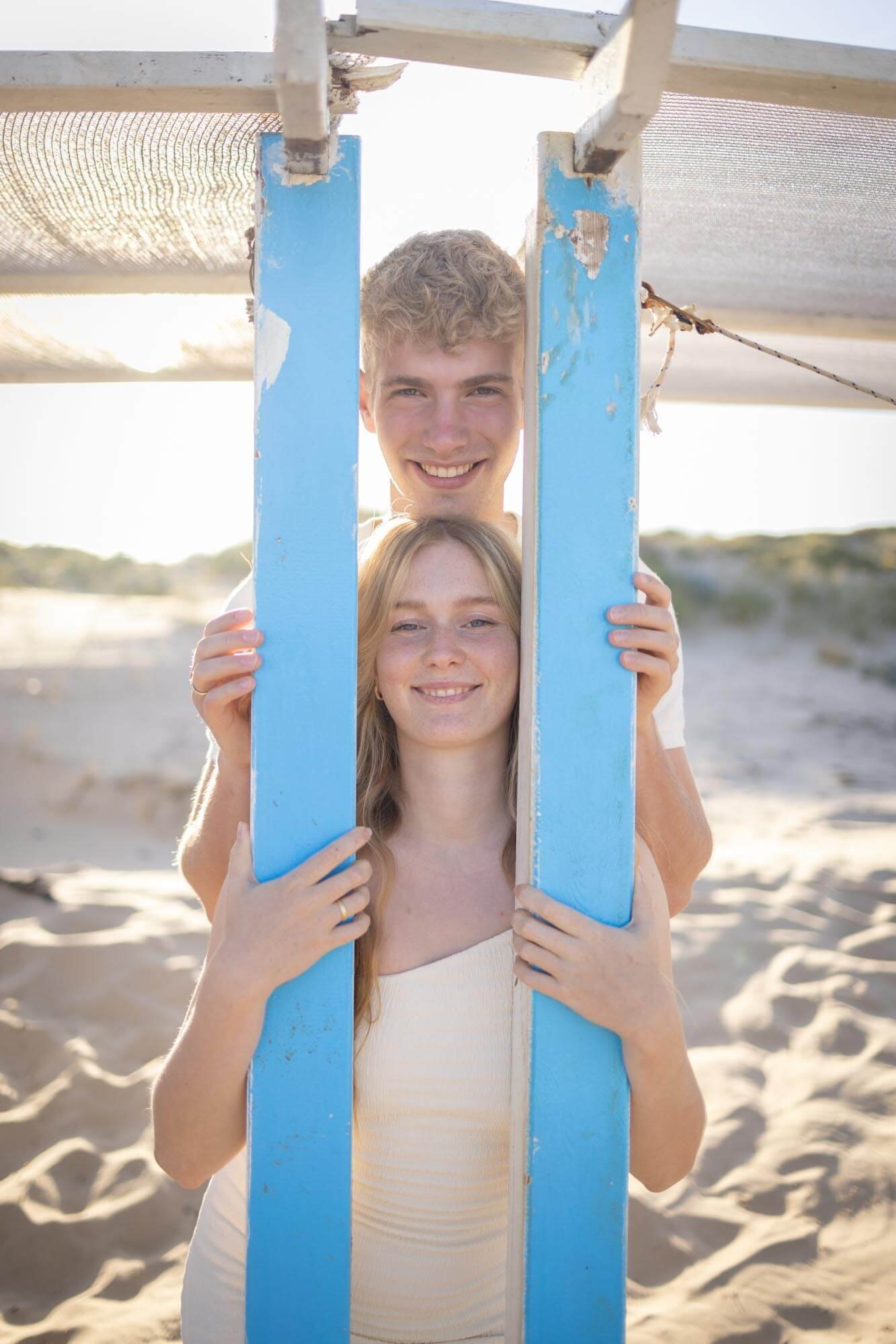 A young man and woman smiling behind a blue wooden frame on a beach.