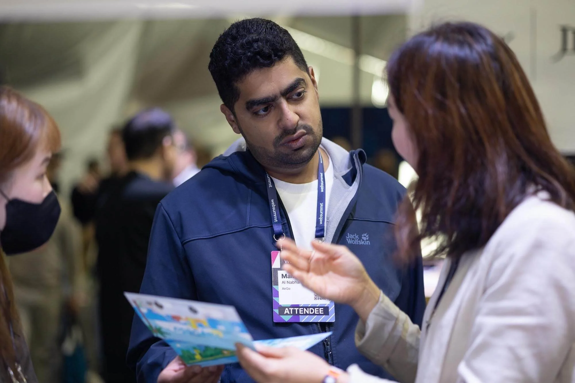 A man with short dark hair and a blue jacket looks attentively at a woman with brown hair, who is explaining something. The woman is wearing a light-colored blazer, and the man has an attendee badge around his neck. People are blurred in the backgrou