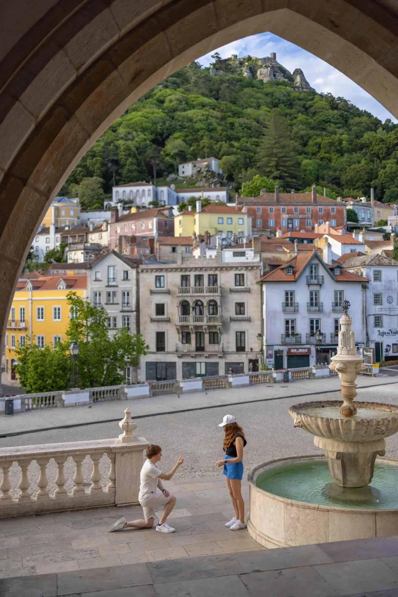 A couple, one kneeling and proposing with a ring, the other standing with a white hat, in front of a fountain on a balcony overlooking colorful buildings and a hilltop castle in a European town.