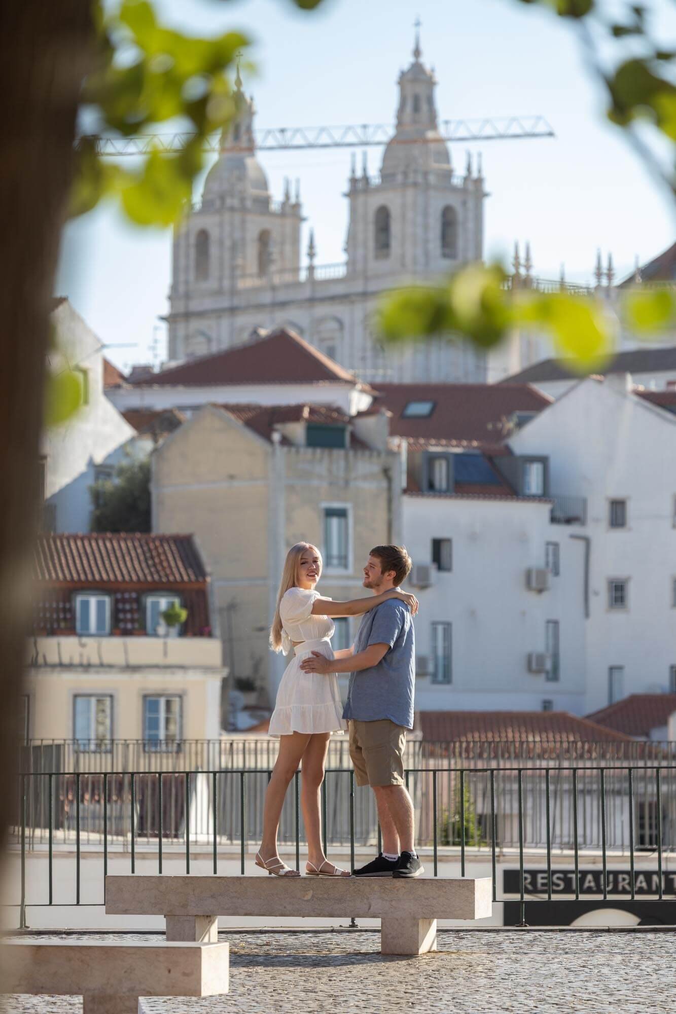A young couple standing on a city bench, embracing and smiling at each other, with old Lisbon church with twin towers in the background, seen through tree foliage.
