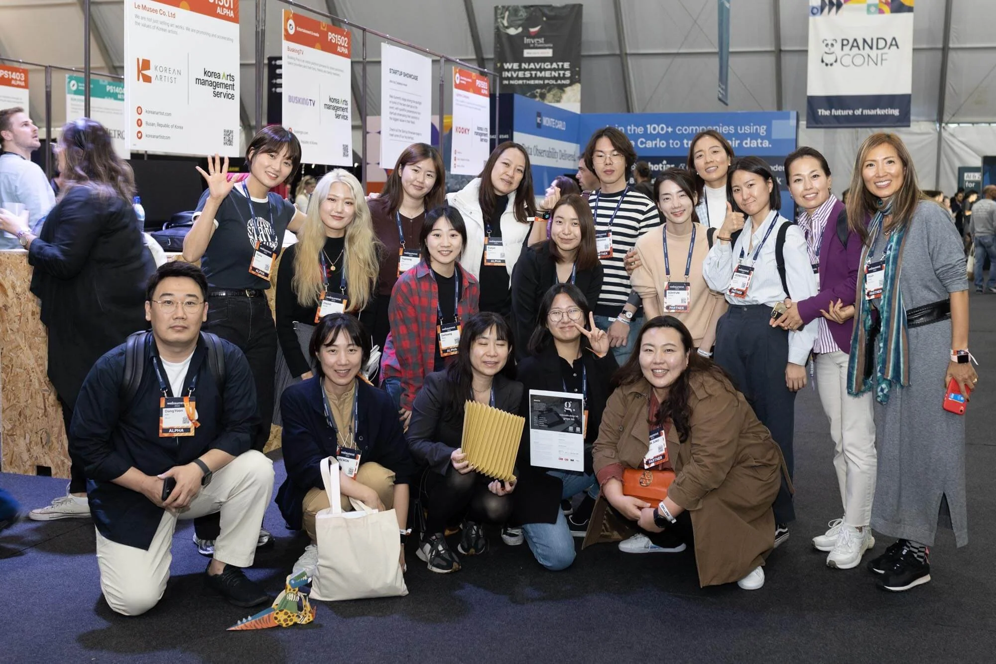 Group of mostly women and two men posing for a photo at a conference or expo, with booths and banners in the background.