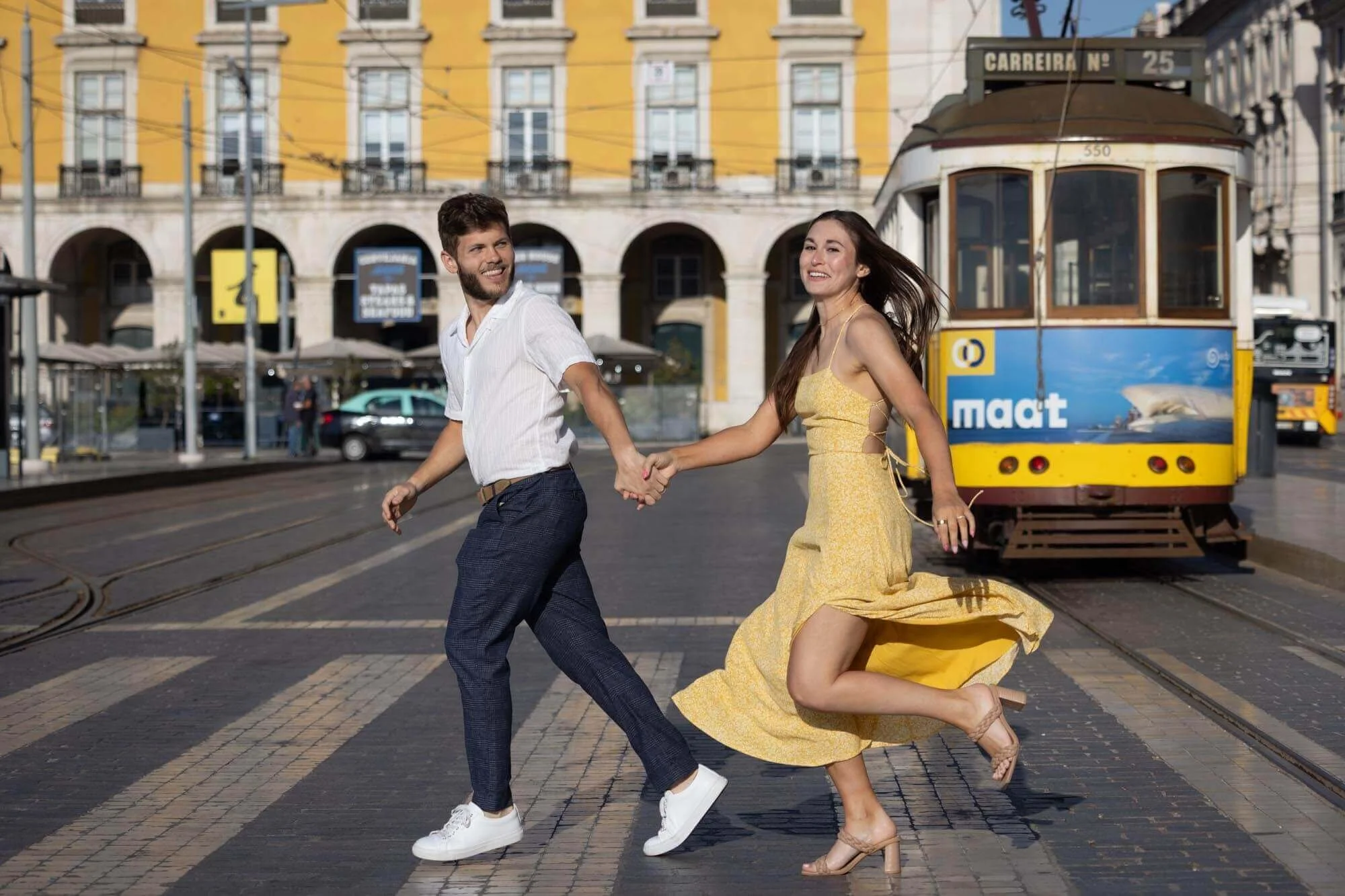A young couple holding hands and dancing on a city street with a yellow tram in the background. The man is wearing a white shirt and dark pants, and the woman is wearing a yellow dress.