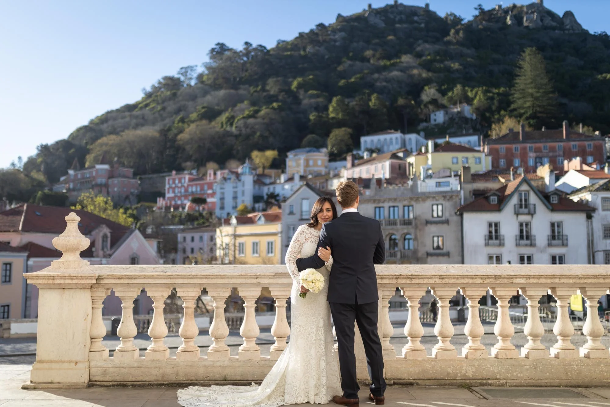 A bride and groom holding hands on a balcony overlooking a colorful hillside town with houses and trees.