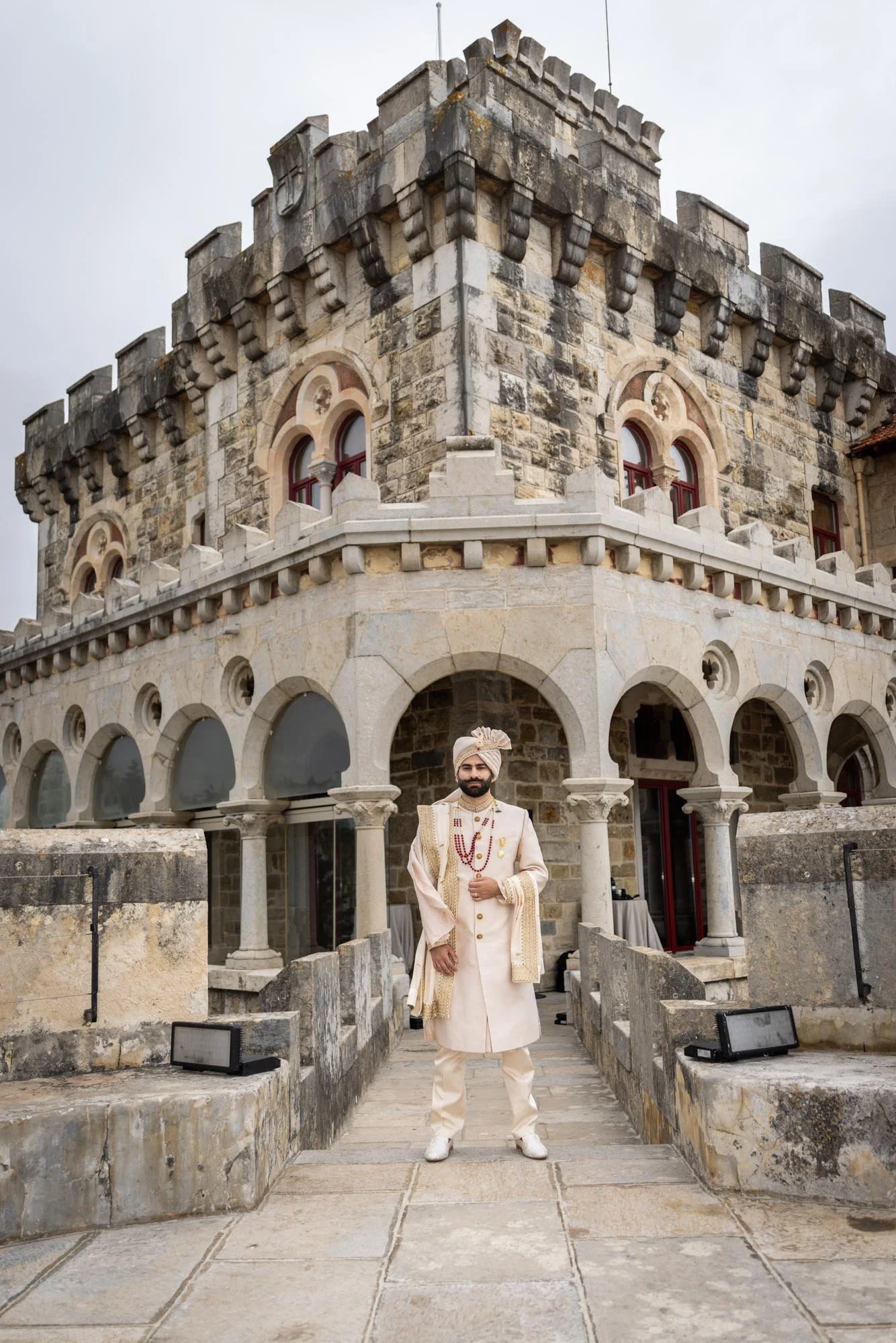 A man dressed in traditional Indian wedding attire standing in front of a historic stone building with arched windows and a battlemented tower.