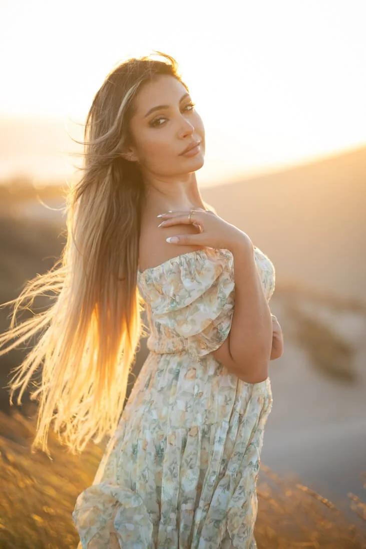A woman with long, blonde hair standing outdoors during sunset, wearing a light floral dress.